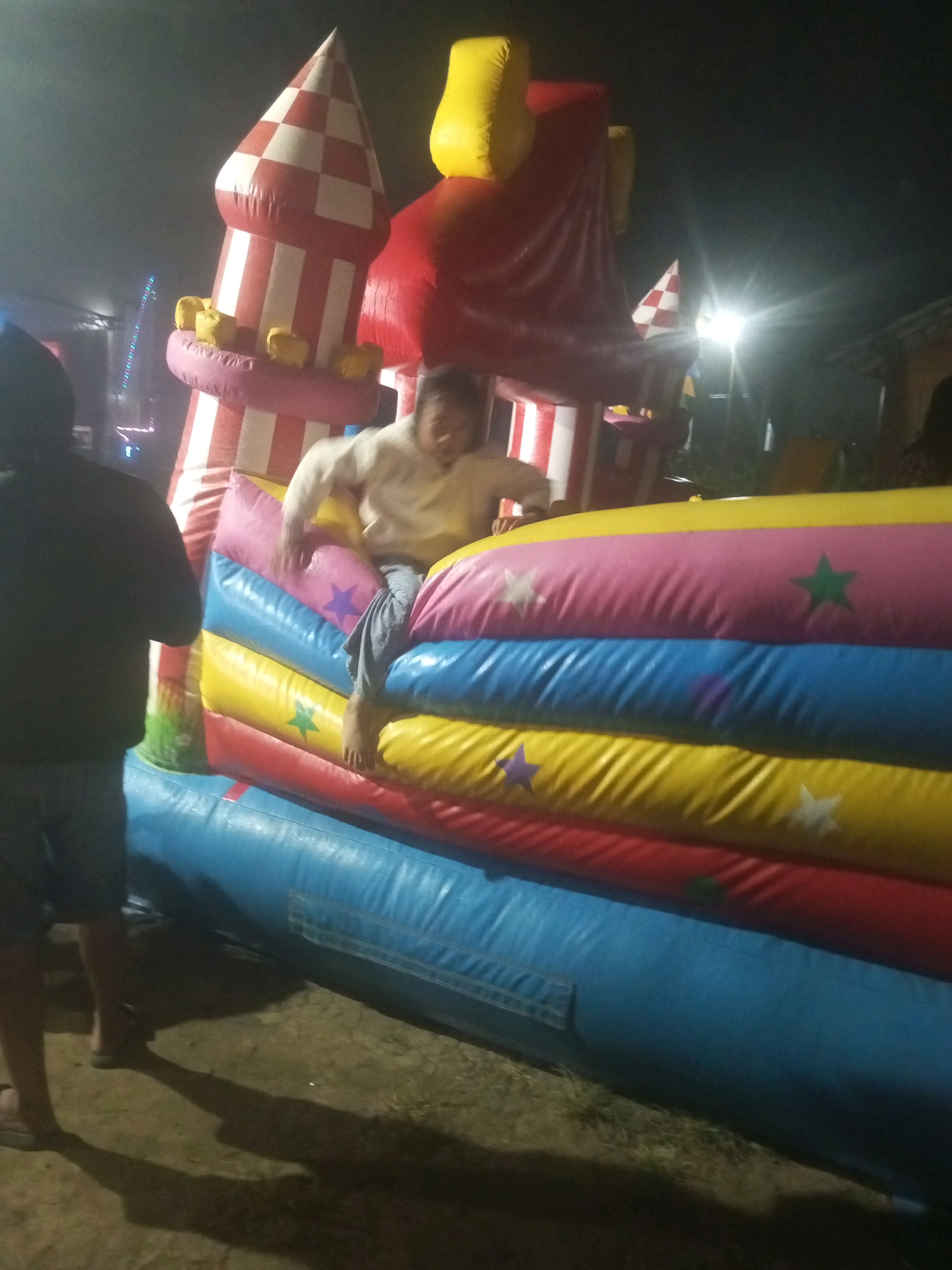The image depicts a nighttime scene at what appears to be an outdoor event in Keseneng, Indonesia. In the foreground, a person, mostly obscured from the back, stands on the left, wearing dark clothing and shorts, with their shadow cast on the ground. To the right, a colorful inflatable castle dominates the frame. A person is actively engaged with the inflatable structure, positioned mid-jump or climb on its multi-colored padded sections. The inflatable is designed with horizontal stripes of yellow, pink, blue, and red, adorned with star patterns in various colors. Behind the inflatable, parts of other structures are visible, including a red and white striped conical tower with yellow accents, and a large red inflatable element. The background is dark, suggesting it is nighttime, with some faint lights and possibly other event infrastructure visible. The overall impression is of a lively atmosphere at a public gathering or fair.