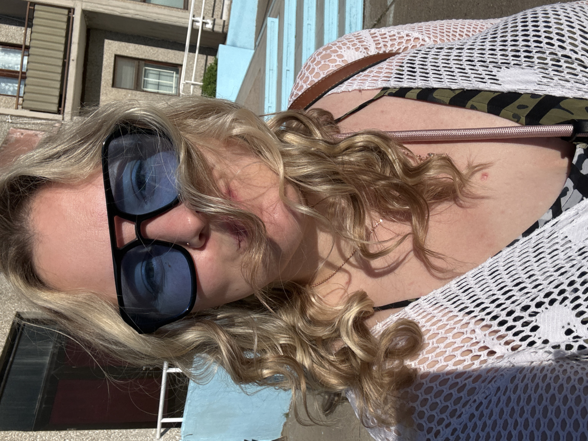 A woman with blond curly hair and sunglasses is lying down outside. She is wearing a white net top over a bikini, and a necklace. A building with windows and a blue fence are visible in the background. The location is unknown and cannot be determined based on the image.