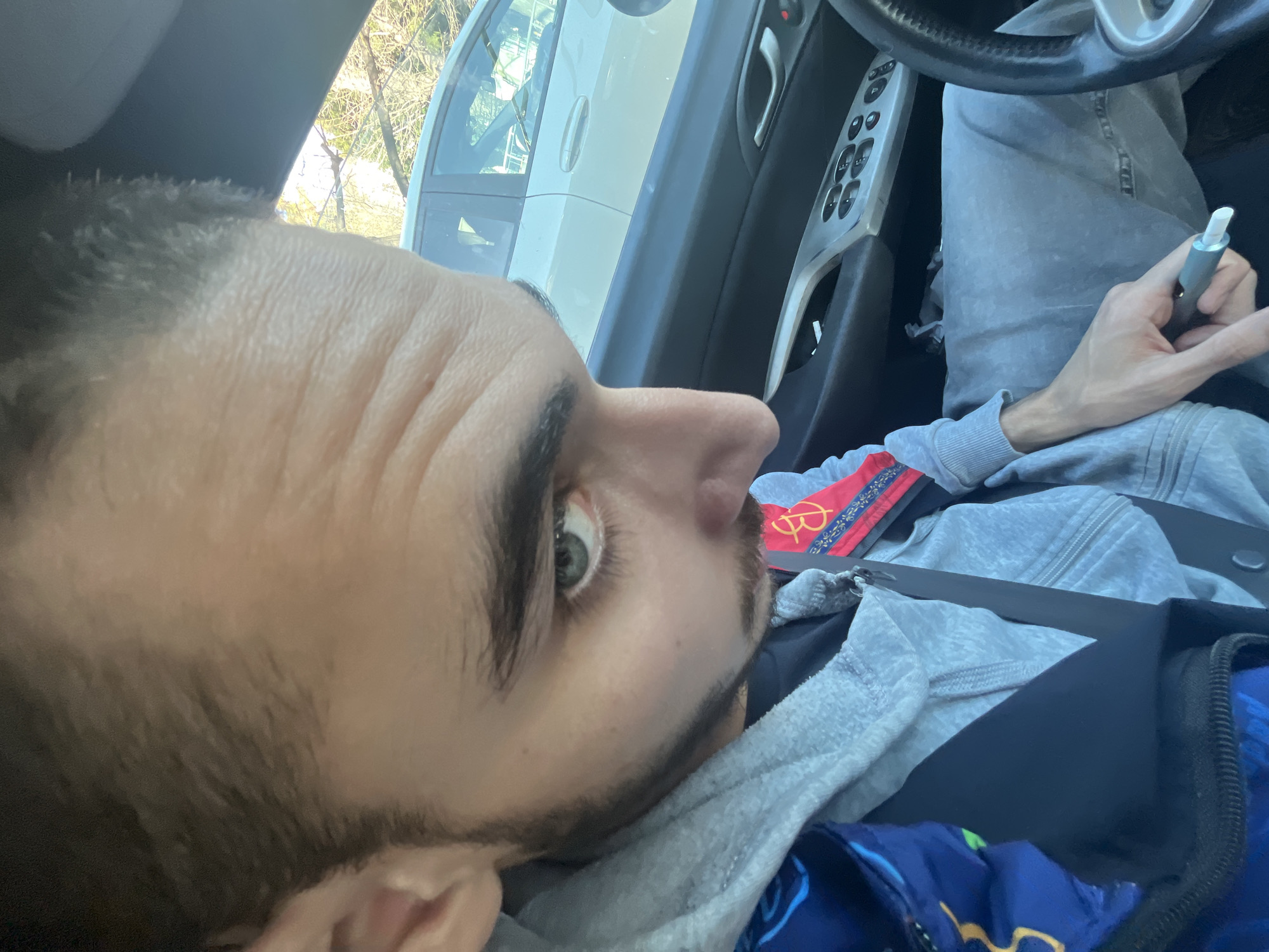 A male individual is observed in the driver's seat of a left-hand drive vehicle, situated in Varna, Bulgaria. He has short dark hair, prominent eyebrows, light-colored eyes, and discernible facial stubble. He is clad in a grey hooded sweatshirt over a darker undergarment, with grey sweatpants. A red fabric bearing a repetitive "B" or "BB" logo is visible on his right side, possibly a bag strap, while a blue item with yellow patterns is partially visible on his left. He is secured by a black seatbelt. His right hand, resting on his lap, holds a two-toned grey and white cylindrical object, consistent with an e-cigarette or vape pen. His gaze is directed downward and to his right. The vehicle's interior is dark-toned, with silver accents on the door panel housing window and lock controls. Through the driver's side window, a light-colored sedan is visible, followed by leafless trees and faint structural elements, indicating an outdoor setting with natural daylight.