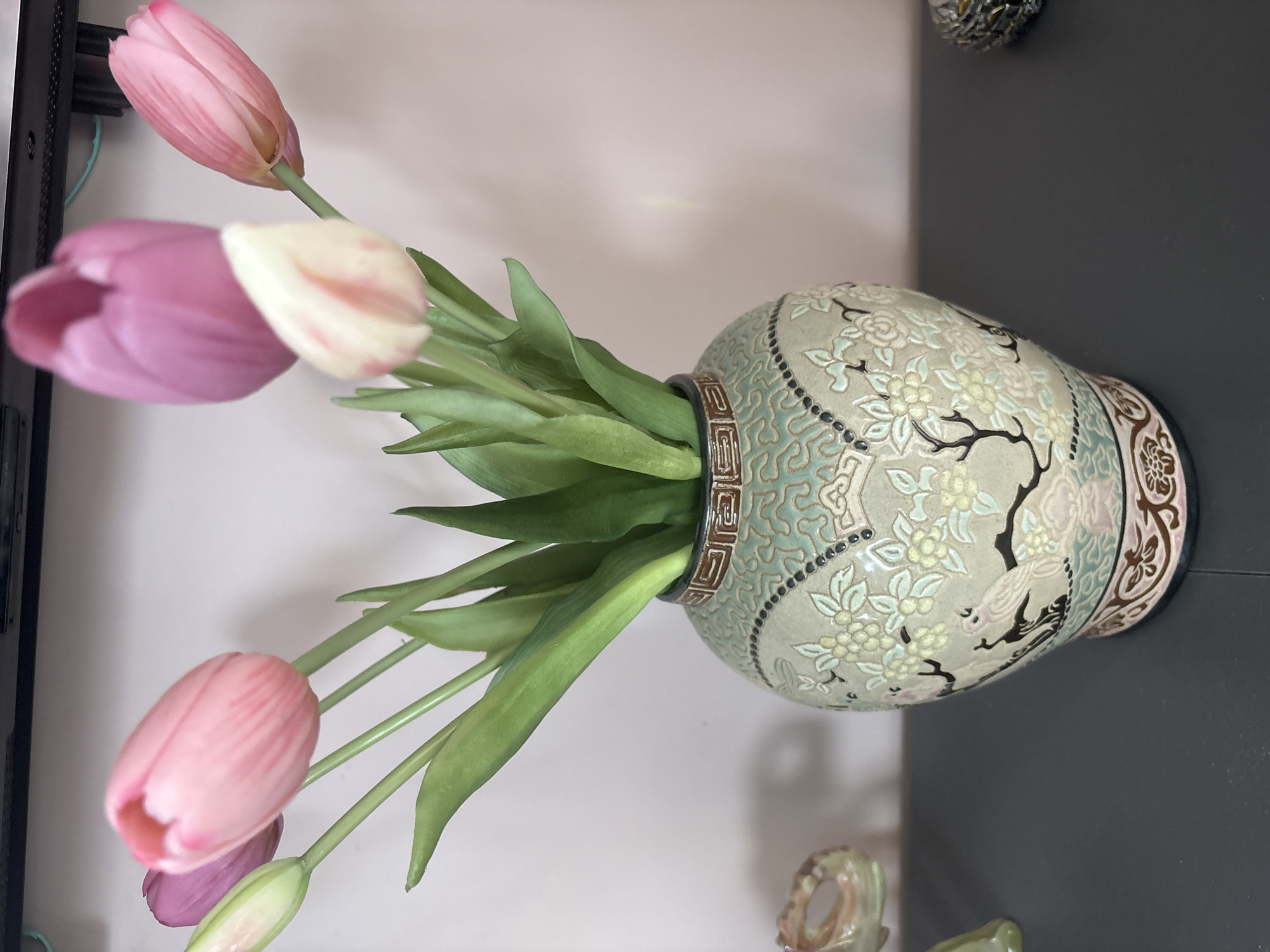 In a dimly lit room, possibly in a residential setting in Dolce Vita KP, Russia, a decorative vase is prominently displayed on a dark surface. The vase, intricately designed with a pale green base, features a detailed pattern of delicate flowers and birds rendered in soft hues of pink, white, and blue. The rim of the vase is adorned with a brown and gold geometric band, adding a touch of elegance. Overflowing from the vase are several tulips, their vibrant pink and white petals contrasting with the muted tones of the vase. The lighting in the room is soft, casting subtle shadows and highlighting the textures of the objects. The background is a plain, light-colored wall, creating a minimalist backdrop that draws attention to the vase and flowers.