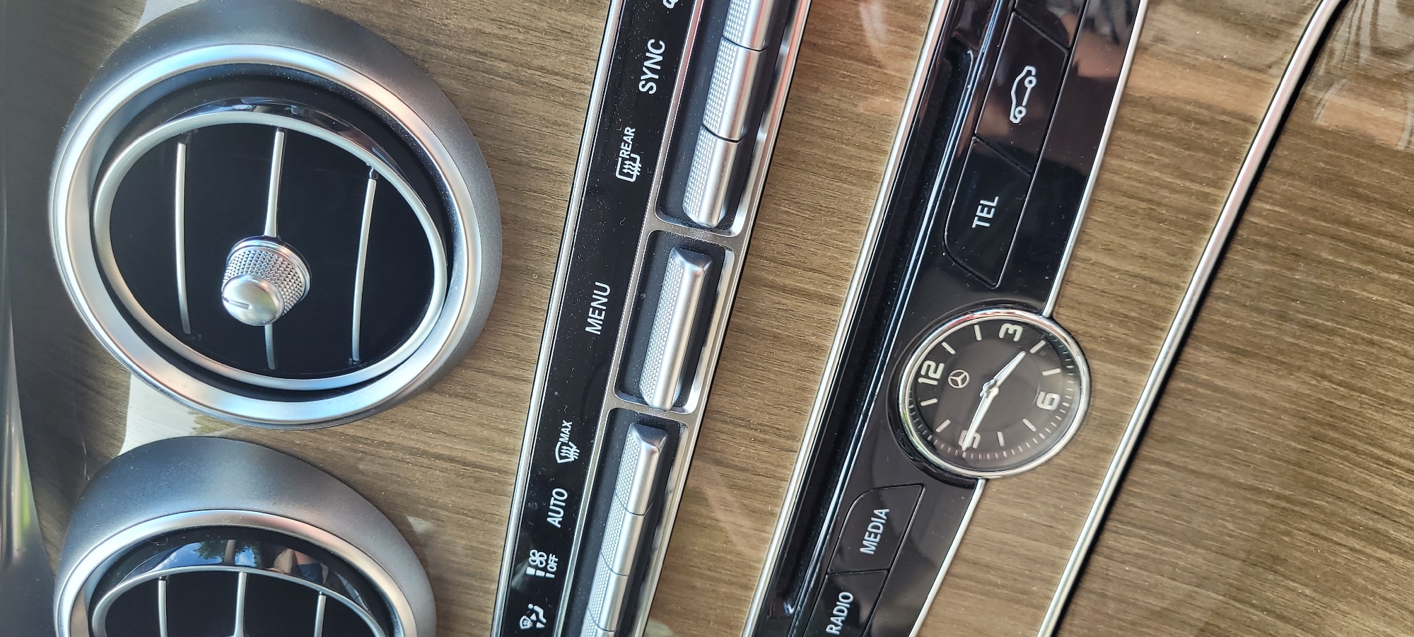 The image shows a close-up view of a car's interior dashboard. Two circular air vents with silver trim are visible at the top. Below the vents are rows of control buttons and a circular analog clock. The controls are labeled with text such as "AUTO," "MENU," "REAR," "SYNC," "RADIO," "MEDIA," and "TEL." The clock face has Roman numerals and a Mercedes-Benz logo. The dashboard has a wood-grain finish. There are no people or animals visible in the image. The scene appears to be indoors, within a vehicle. The image does not provide any information that would allow for its location to be identified as Riga, Latvia.