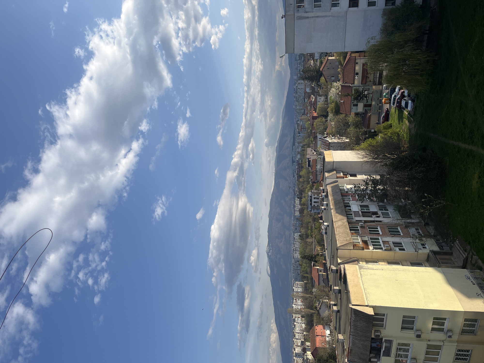 A vertically oriented photograph captures a cityscape with a mountain range in the background, likely taken in Sofia, Bulgaria. The sky, predominantly blue, is filled with scattered cumulus clouds. A series of residential buildings and houses are visible, with a mix of architectural styles and colors, including red-tiled roofs and yellow facades. Some cars are parked on the side of a patch of grass, in front of the buildings. In the foreground, buildings of varying heights are seen, suggesting an urban environment. Behind the city, a mountain range is visible, partially obscured by clouds. A black cable is visible in the upper left corner.