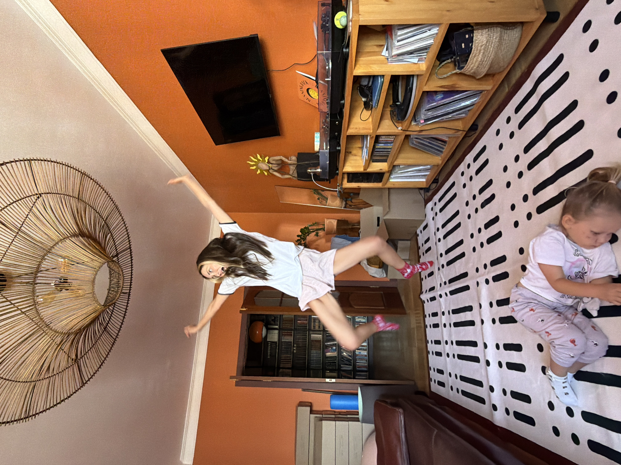 This indoor photograph, taken in Khimki, Russia, captures a lively scene featuring two girls in what appears to be a living room or recreational space. The room is characterized by a bright orange wall on the right and a lighter, possibly white, ceiling and wall on the left.
The primary subject is an older girl, positioned centrally, mid-jump or strike a playful pose with her arms outstretched and legs bent. She wears a white t-shirt with dark trim and light-colored shorts, along with patterned pink socks. Her hair is long and dark with lighter streaks, and her expression suggests excitement with an open mouth and wide eyes. To her right, a younger girl lies on a distinctive white rug patterned with black dashes and dots. She wears light-colored pajamas and appears focused on something in her hands, her expression calm.
The room is furnished with a variety of objects. A large, intricate circular woven chandelier hangs from the ceiling on the far left. On the orange wall, a flat-screen television is mounted. Below the TV, a tall, light-wood shelving unit is packed with numerous CDs or vinyl records. A unique decorative item resembling a sun-headed figure is also visible on a lower shelf of this unit. In the background, a doorway reveals another room with bookshelves. A brown leather sofa or armchair is partially visible in the bottom right corner.
The lighting suggests it is daytime. On one of the shelves, the word "MAGNAVOX" is visible on what appears to be a book or album cover. The overall atmosphere is casual and domestic, with an energetic quality lent by the older girl's pose.