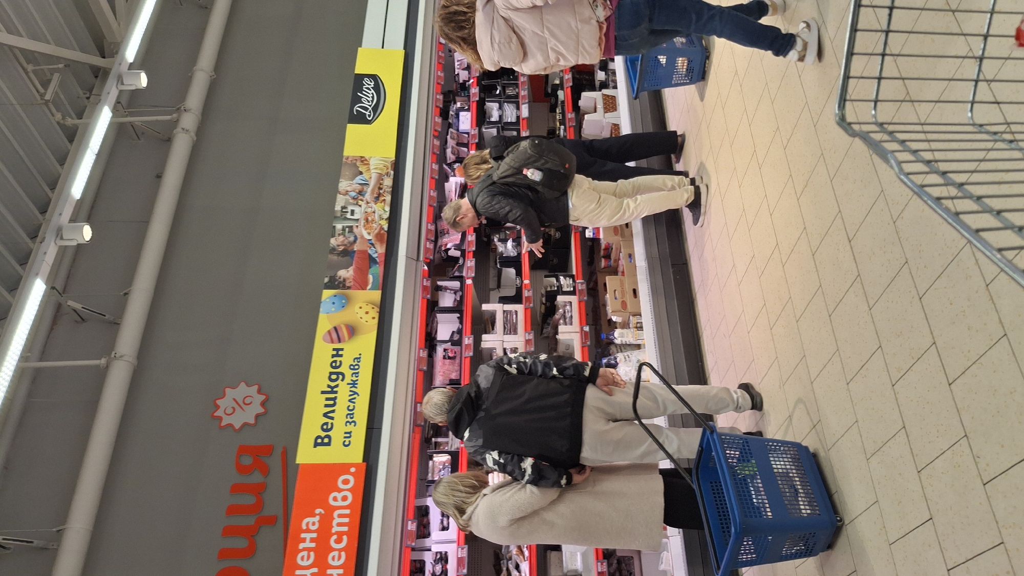 People are shopping in a supermarket. The shoppers are near a refrigerated section displaying various products. There is a shopping cart nearby. The supermarket's interior features tiled flooring and bright lighting, with signage in Bulgarian visible on the walls.