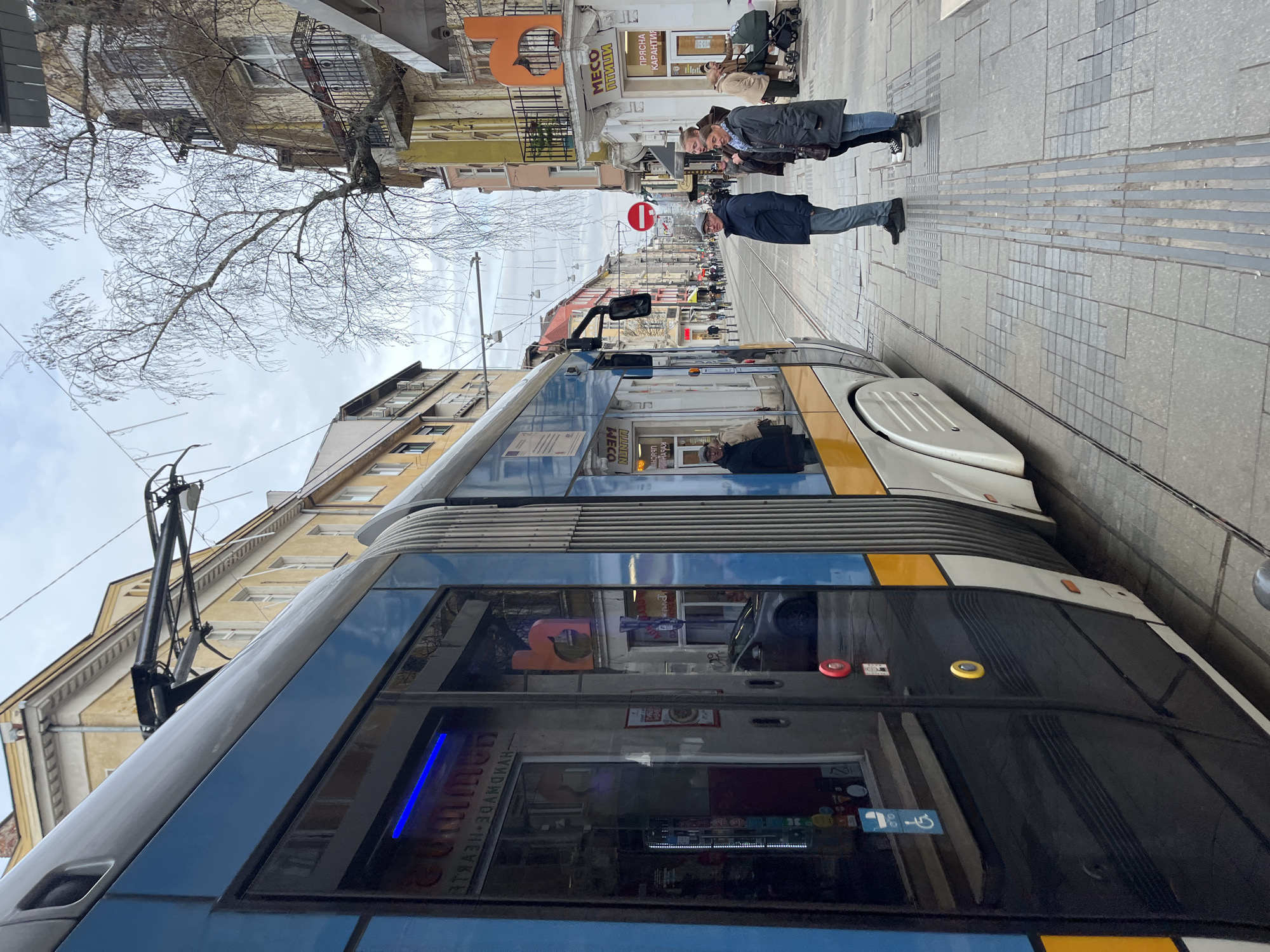 The image shows a street scene in Sofia, Bulgaria, with a modern tram occupying much of the foreground. The tram is predominantly blue, black, and white with yellow accents. The tram's windows reflect the surrounding buildings, including shops with signage in Bulgarian.
In the background, a street extends into the distance, flanked by buildings of varying heights and architectural styles. Pedestrians, including a couple, walk along the paved sidewalk. A "no entry" road sign is visible in the distance. The sky is overcast, and bare branches of a tree are visible near the top left corner.