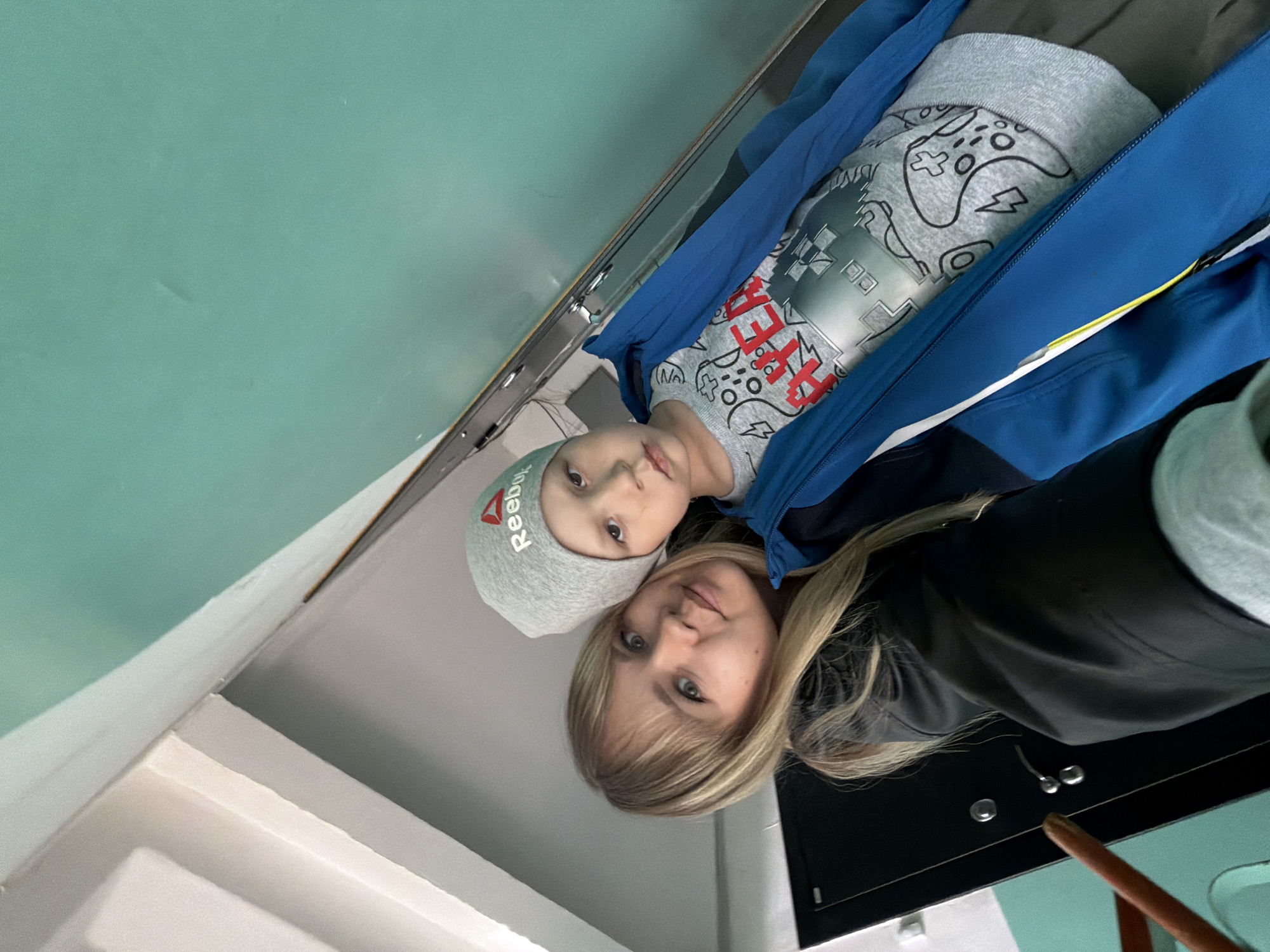 This image features an adult female and a young male child, likely a mother and son, looking directly at the camera from a low, tilted angle. The setting is indoors, with a prominent light teal or mint green wall in the upper half of the frame. A white or light gray wall/ledge is visible in the lower left, and dark surfaces with metallic fixtures are in the lower right, suggesting a modern interior.
The adult female has long blonde hair with a fringe and light eyes, wearing a dark jacket. The young boy, positioned in front of her, has light brown hair and light eyes. He is wearing a gray knitted beanie with the "Reebok" logo in green text. His gray long-sleeve shirt displays a graphic design of game controllers and the word "HYPER" in bold red text. He is also dressed in a blue jacket with a yellow accent stripe. Both individuals have neutral expressions, calmly posing for the camera. The lighting is soft and even, typical of an indoor environment. The location is Chaikovsky, Russia.