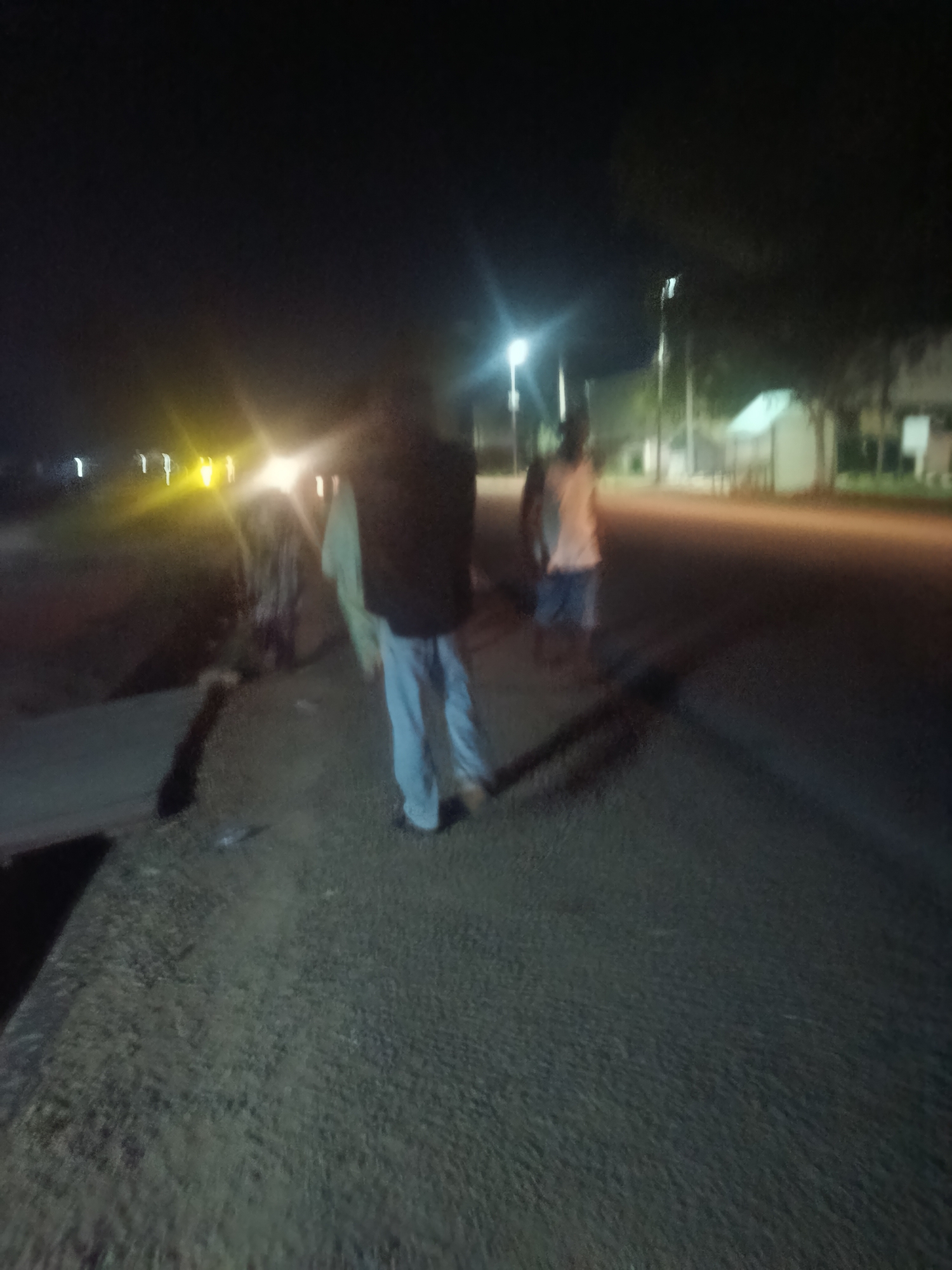 The image depicts a dimly lit, outdoor scene at night, likely in Jalingo, Nigeria. Several individuals are visible, appearing as blurry figures due to the low light conditions and possibly motion blur.
In the foreground, a person wearing light-colored pants and a dark vest or jacket is walking away from the camera. To their left, another figure is partially visible, possibly carrying or wearing a light-colored garment. Further back, two other figures are visible; one appears to be wearing a white tank top and dark shorts, and another is more obscured.
The environment suggests an urban or semi-urban setting. Streetlights are present, casting streaks of light across the scene. A road surface is visible, appearing to be asphalt or packed earth. In the background, there are indications of buildings and trees, though they are indistinct due to the darkness. The overall impression is a snapshot of people moving through a town or city at night.