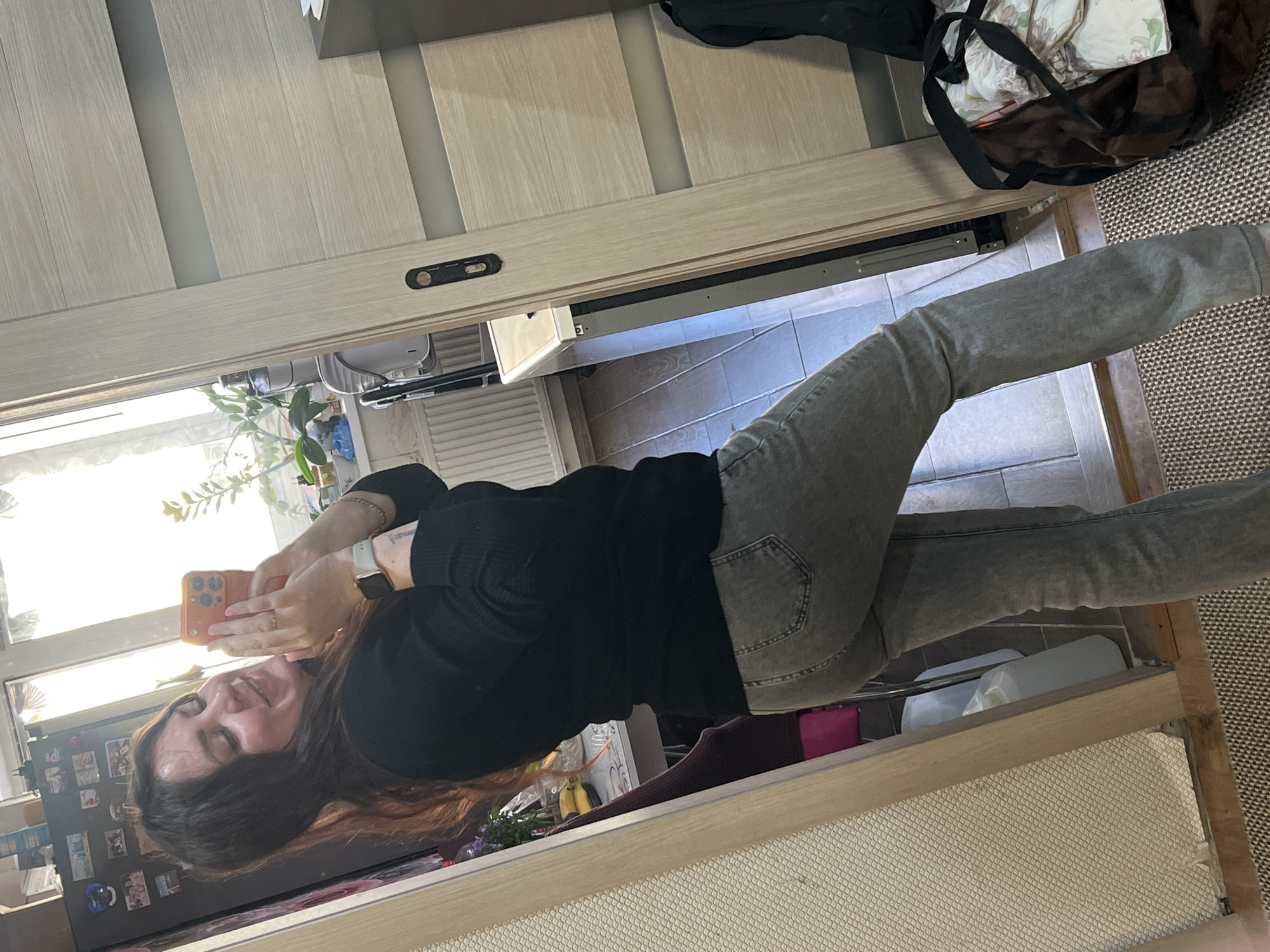 A woman is standing inside a building taking a selfie. The woman has long brown hair, is wearing a black long-sleeved shirt, gray jeans, and an Apple watch on her left wrist. She is holding an orange iPhone in her hands.
Behind her is a refrigerator with pictures on it. There are flowers and bananas on a table near the refrigerator. She is standing near a window.
The location is Noviy, Russia.