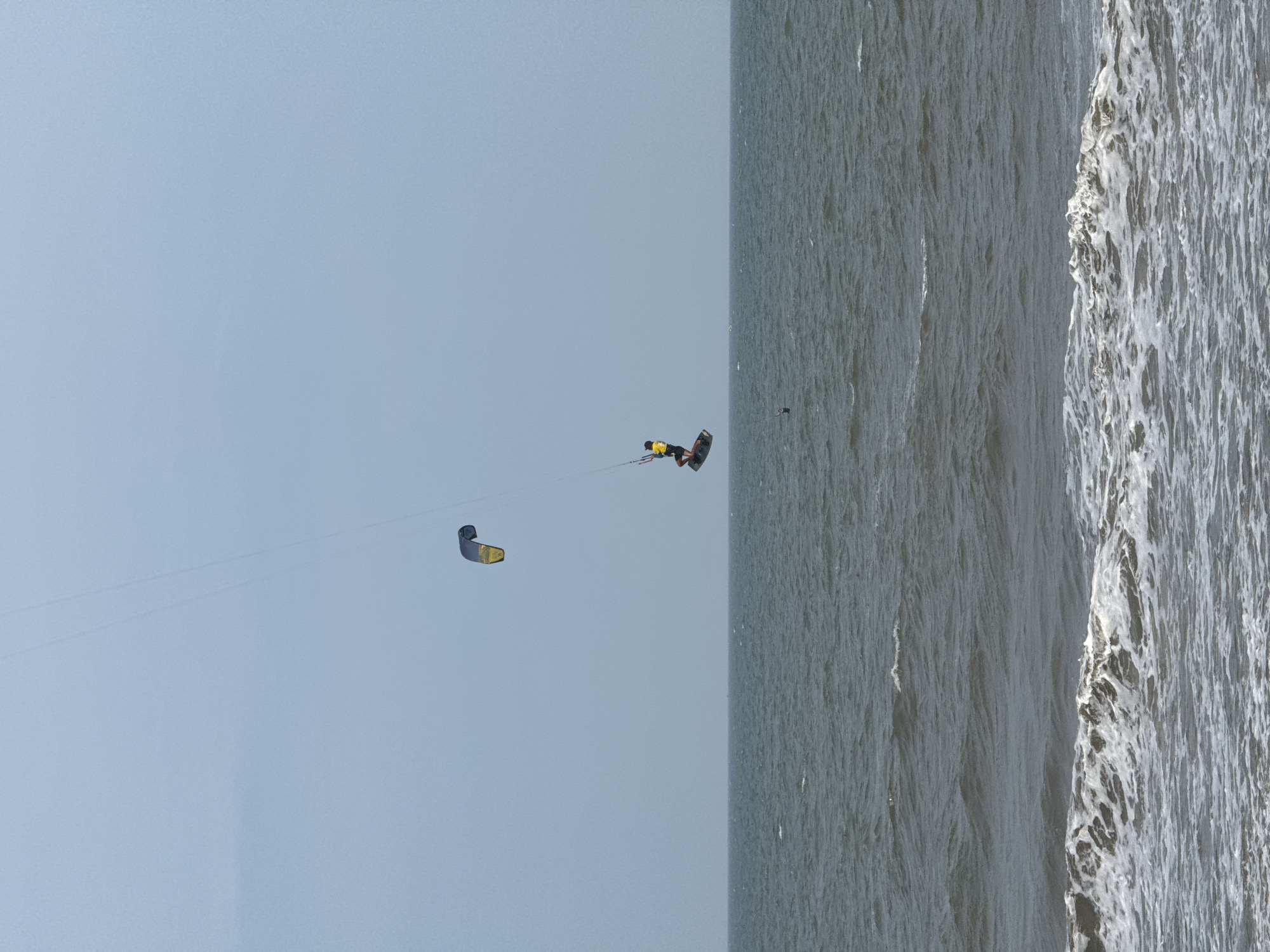An aerial shot captures two kitesurfers in action over the sea, in Phan Thiết, Vietnam. The kitesurfers are positioned at different heights, with one actively riding a board and suspended in mid-air, while the other is higher up and seemingly still. The sea is choppy, with white-capped waves indicating moderate conditions. The sky is overcast, casting a diffused light over the scene. A faint line runs from the upper left of the frame downwards, suggesting the presence of additional kites or support lines not fully visible. A small, distant figure can be seen in the water, possibly another kitesurfer or swimmer. The image is a still shot, freezing a dynamic moment of watersport against a serene, yet energetic, coastal backdrop.