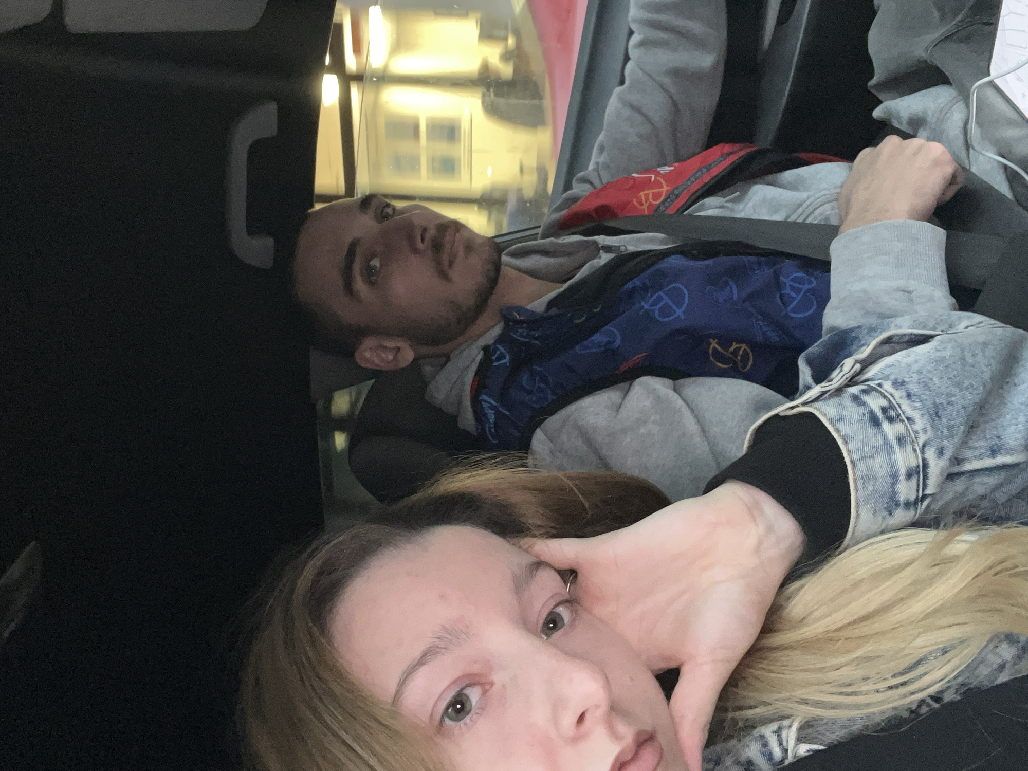 Two individuals are positioned inside a vehicle, captured from a selfie perspective originating from the passenger side. In the foreground on the left, a light-skinned female with long blonde hair and green-brown eyes looks directly towards the camera, her left hand resting against her jawline. She is dressed in a light blue denim jacket over a dark long-sleeved garment. To her right, in the driver's seat, a light-skinned male with short dark hair and visible stubble on his face looks slightly to the right of the camera. He wears a grey hooded sweatshirt overlaid with a blue and red patterned vest or scarf, and his seatbelt is visibly fastened across his chest. The vehicle's interior includes a dark headliner and a passenger grab handle above the male subject. Through the driver's side window, artificial lighting from an external building with rectangular windows is visible, along with a partial view of a red vehicle. The lighting conditions suggest dusk or nighttime. The specific location within Varna, Bulgaria, cannot be ascertained from the visual evidence presented.
