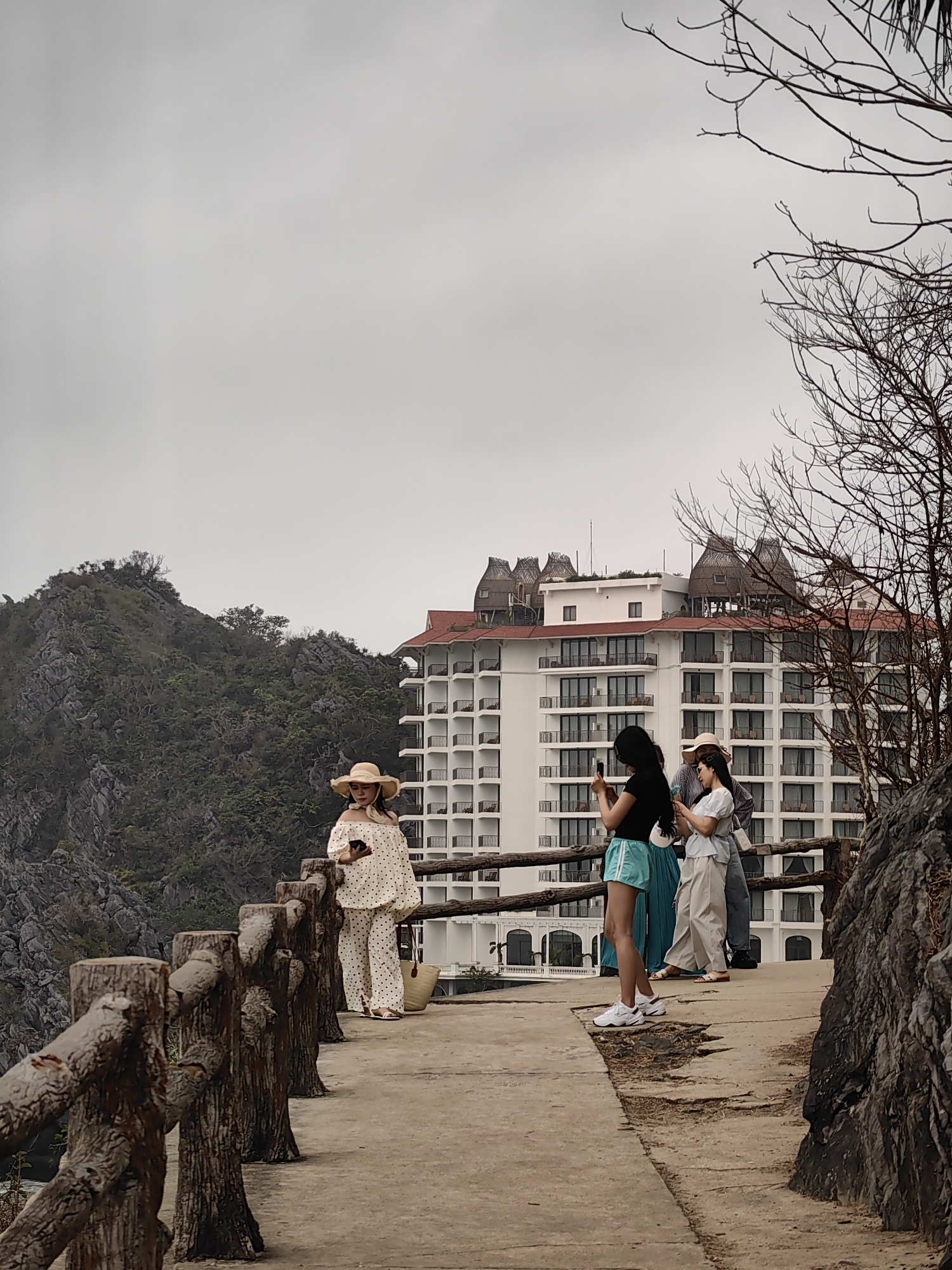 On an overcast day in Hải Phòng, Vietnam, four people stand on a concrete path bordered by a rustic wooden fence, appearing to be a walkway. The path is cracked and shows signs of wear.
To the left, a woman in a polka-dot off-the-shoulder blouse and matching pants, topped with a straw hat, leans against the fence, looking at her mobile phone. A straw bag rests near her.
Further along the path, three other people are present. One woman, dressed in a black top and blue shorts, holds her phone up, apparently taking a picture. A second woman, dressed in light-colored clothing and also wearing a hat, appears to be looking at her phone as well. A person dressed in darker clothing stands next to them.
Behind the group is a large, multi-story hotel or resort building with a distinctive red roof and white exterior. Some of the rooms have balconies. To the left of the building is a rocky, tree-covered mountain. To the right of the group is a rocky outcropping with bare tree branches. The sky is uniformly grey and overcast.