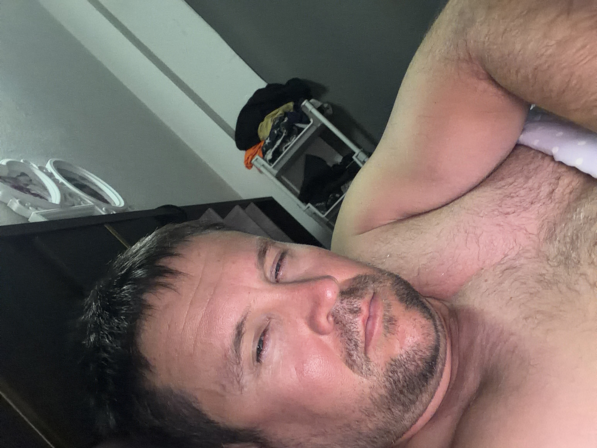 The image is a close-up selfie of a man lying down, with his head tilted and his right eye partially closed. He has short, dark hair and stubble on his face. His skin appears flushed. In the background, a shelf unit with clothing is visible, as well as a framed picture on a dark surface. The lighting is dim. The image does not provide any information about the location being Baan Mae Nam, Thailand.