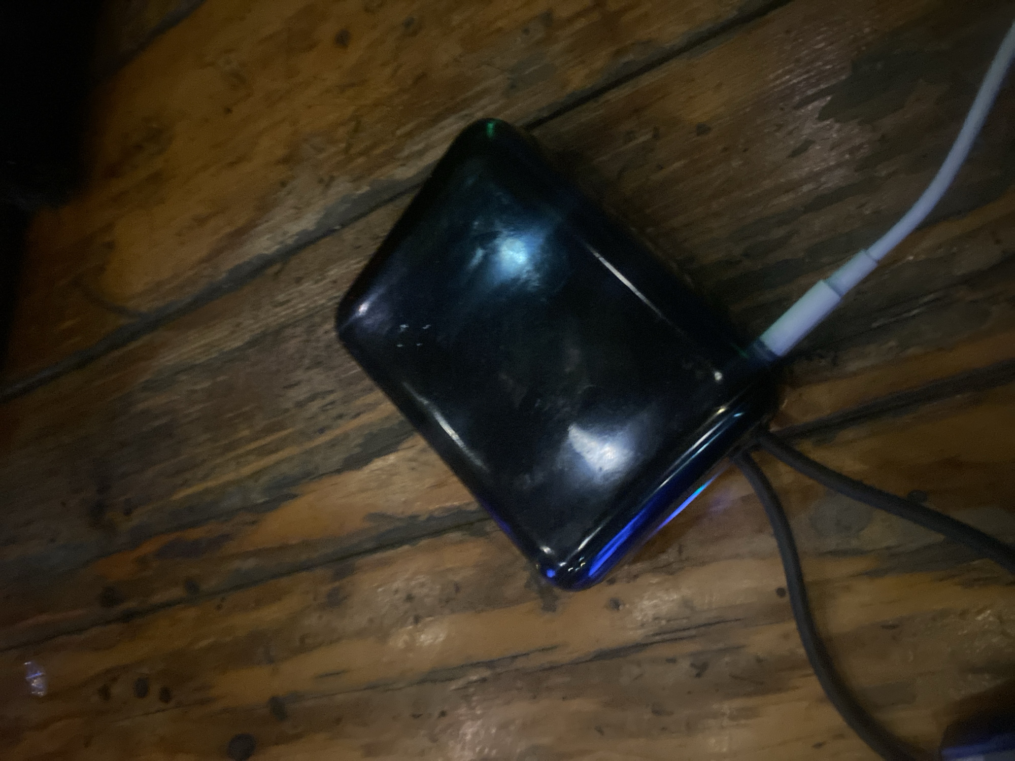 The scene is located in the city of Howayi, Nigeria. A dark, glossy, rectangular electronic device with rounded corners, possibly a power bank or portable hard drive, rests on a wooden surface. The device displays a prominent blue light on its upper left corner and a small green indicator light on its upper right. A light-colored cable, appearing white or light gray, is connected to the device on its lower right side, extending out of frame. A second, darker cable, likely black, is partially visible beneath the device, extending towards the lower left. The surface supporting the device consists of distinct wooden planks or panels, exhibiting varying shades of brown, visible grain patterns, and darker streaks, suggesting a textured, possibly rustic, wooden table or floor. The ambient lighting is subdued, highlighting the illuminated indicators on the device. No people or human interactions are visible.