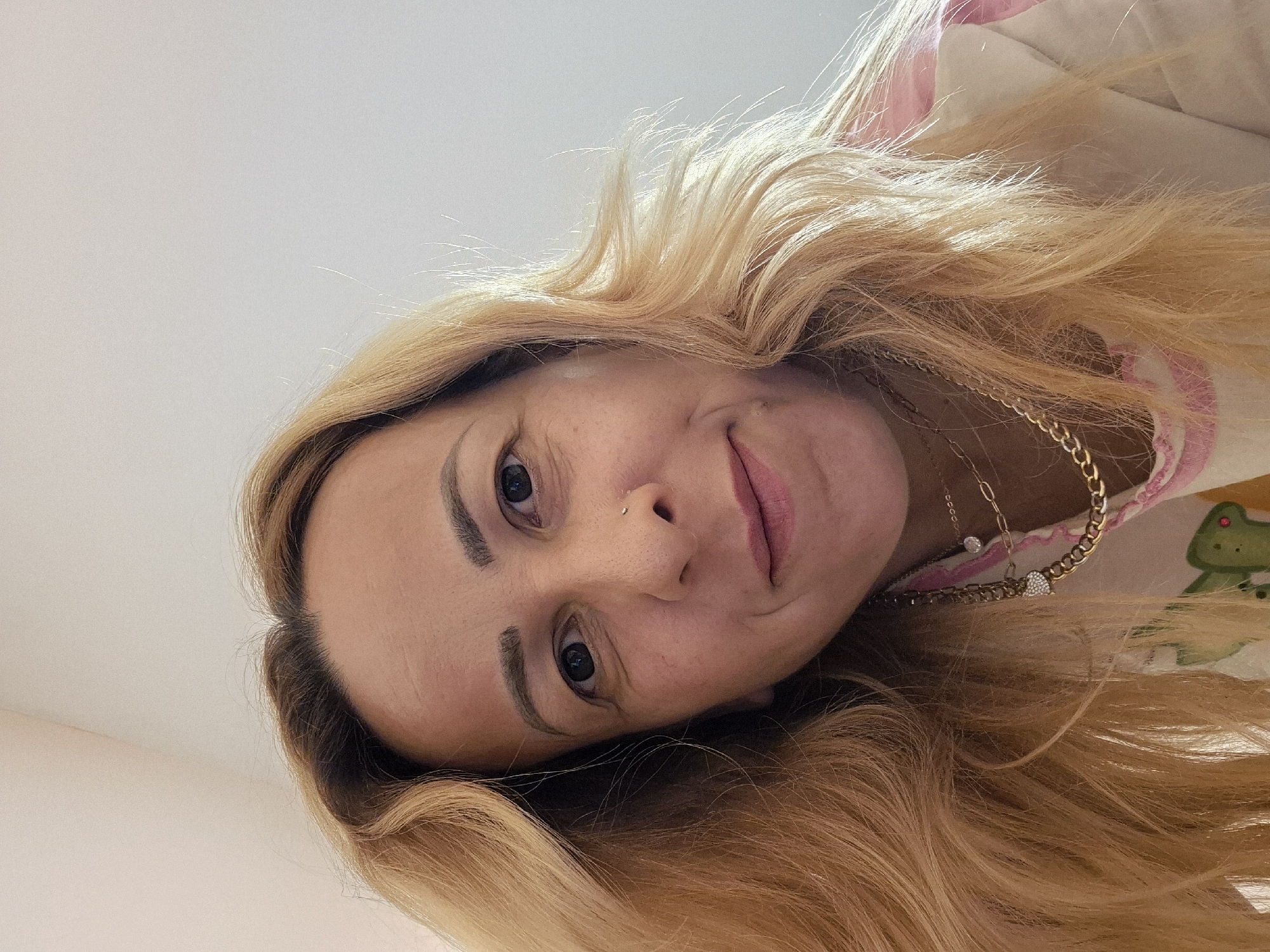 A woman with blonde hair is looking up at the camera. She is wearing a pink and white shirt with a green design. She is also wearing two gold necklaces. The background is a plain white wall. The image was taken indoors, in what appears to be a home environment. There are no other people or objects of note in the image. The lighting is natural, coming from an unseen source to the left of the frame. The woman's expression is neutral, and she is not actively engaged in any particular action. The overall tone of the image is casual and personal.
