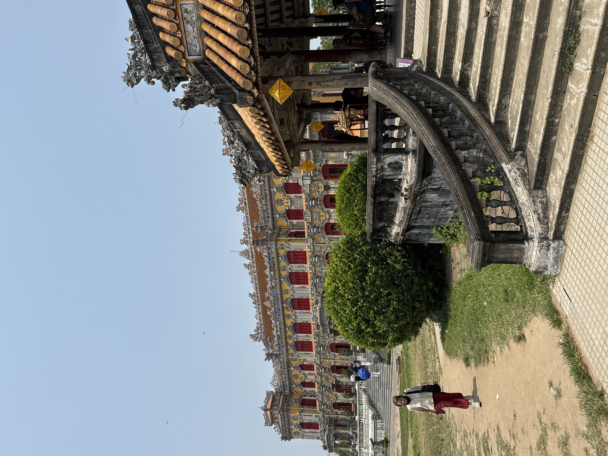 The image depicts a building in Phú Xuân Ward, Vietnam. The building has a tile roof with yellow and orange accents, red windows, and ornate carvings. In front of the building, there is a paved walkway, a dirt path, and a grassy area with a round bush. A person in a light shirt and red pants is standing on the dirt path. To the right of the building, a staircase ascends. The sky is clear blue.