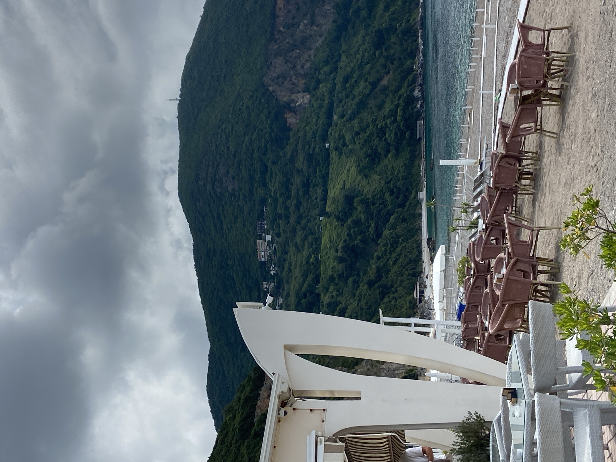 The image depicts an outdoor seating area in what appears to be a beach or coastal setting in Prijevor, Montenegro. Dominating the background is a large, green, densely forested mountain under a cloudy sky. In the midground, there is a beach with rows of brown plastic chairs and white lounge chairs, some arranged around glass-topped tables. The sea is visible as well, with clear turquoise water. In the foreground, there is a white structure, possibly part of a building or canopy, with curved architectural elements. No individuals are clearly visible, though there is a partially obscured figure near the base of the structure. The environment suggests a tourist or recreational area on a partly overcast day.