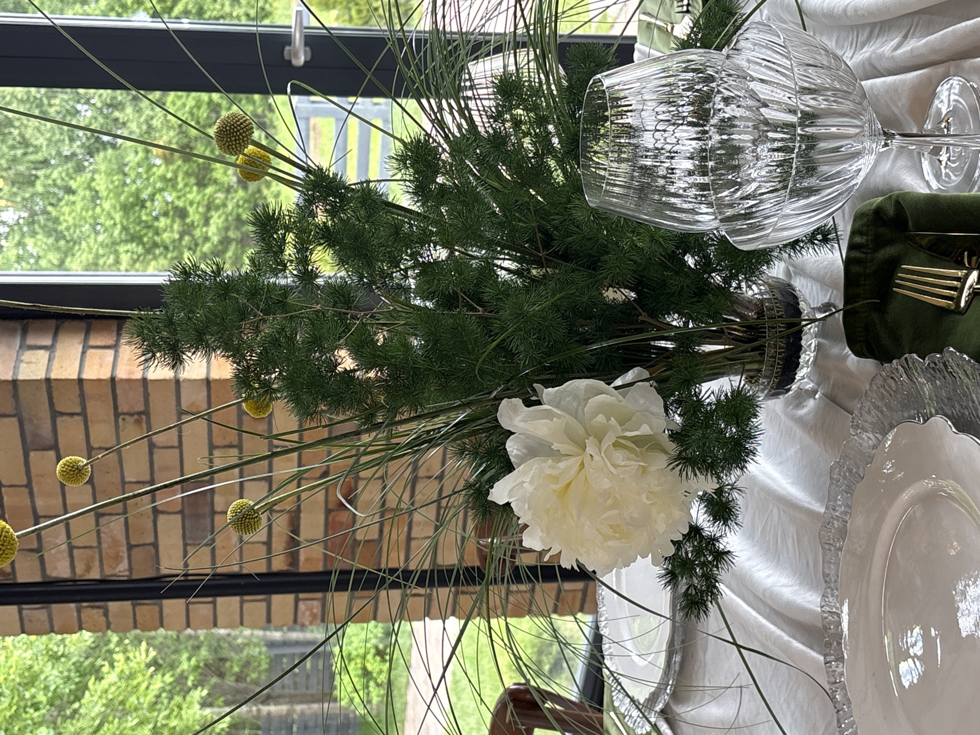 The image displays an indoor dining setting, likely a restaurant or event space, with a table prepared for guests. The central element is a large floral arrangement in a clear glass vase, featuring a prominent white peony at the base, surrounded by dark green, spiky foliage resembling conifer branches, and long, thin, grass-like green stalks. Scattered among the greenery and extending upwards are several spherical, textured yellow flowers on slender stems.
The table is draped with a white or light-colored tablecloth exhibiting soft folds and a subtle sheen. Partially visible on the left is a place setting, including a clear glass charger plate with a ruffled edge, topped by a white dinner plate with a gently undulating rim. On the right side of the table, two clear glass goblets with distinct vertical fluting are stacked, suggesting a wine glass above a wider, shallower coupe. A dark green cloth napkin, folded, rests beneath the glassware, with a gold-colored fork and knife tucked into its folds.
The background reveals a large window or glass door, through which a dense expanse of lush green trees and foliage is visible, indicating an outdoor natural environment. Part of an interior brick column with reddish-brown bricks and lighter mortar is visible behind the floral arrangement on the left. A dark, possibly black, frame borders the window/door, and a silver-colored door handle is discernible on the right side of the frame. No individuals are present in the scene, and no specific actions are depicted. The image itself provides no visual evidence to determine its geographical location as Perm, Russia.