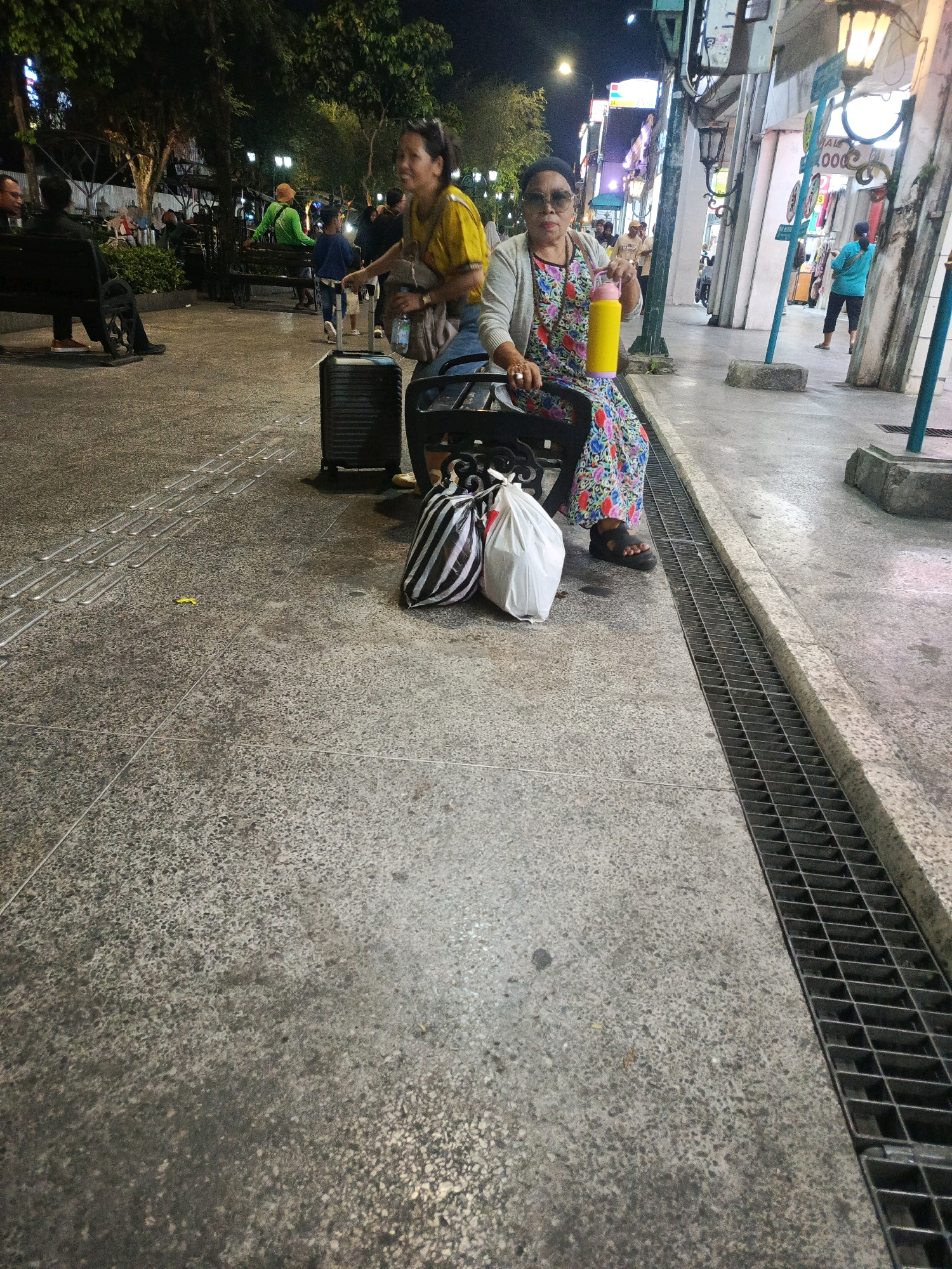 An evening scene captures a pedestrian area in Yogyakarta, Indonesia, likely Malioboro Street, given the bustling atmosphere and the distinct architectural style of the buildings lining the thoroughfare. Two women are the central figures. The woman in the foreground, seated on a black bench, wears sunglasses, a light-colored cardigan over a colorful floral dress, and dark sandals. She holds a yellow water bottle in her right hand. Beside her, on the ground, are two bags: one with black and white stripes and a plain white bag. A black suitcase stands behind her. To her left, another woman in a yellow top is visible, appearing to be in motion, with a handbag slung over her shoulder and a water bottle in her hand. In the background, other pedestrians are seen, some seated on benches, and streetlights illuminate the area, casting a warm glow on the scene. The ground is paved with textured concrete, featuring tactile paving for the visually impaired. A metal grate runs along the edge of the walkway, suggesting a drainage system. Trees are present, adding to the urban greenery. The overall impression is one of a lively public space at dusk, with people enjoying the evening.