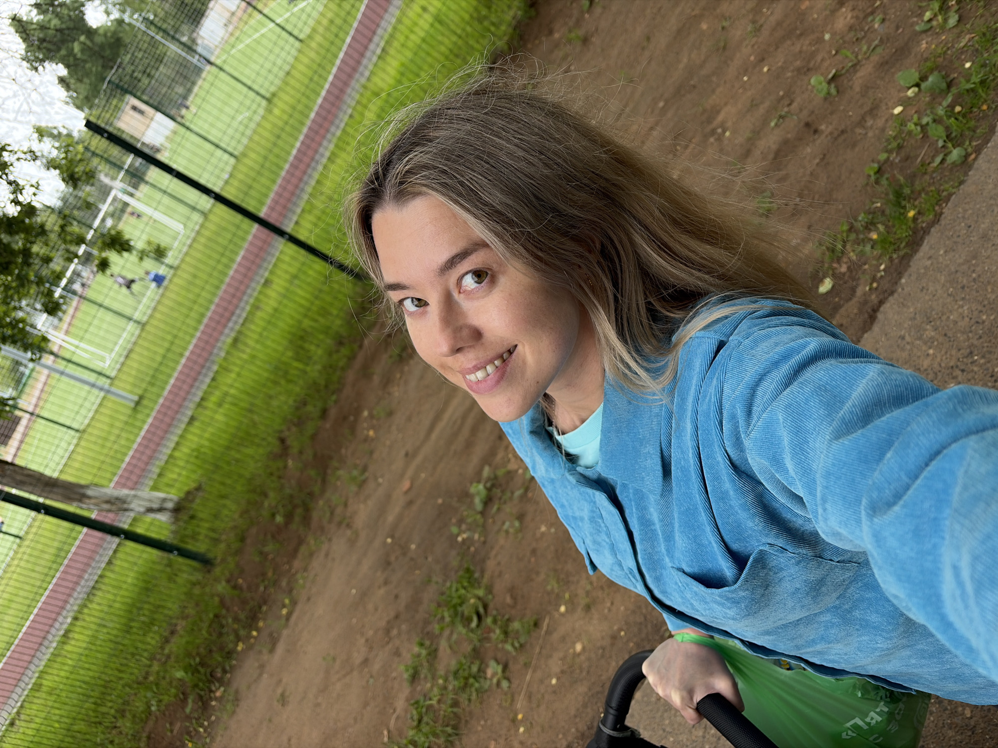 A young adult woman takes a high-angle selfie in Chaikovsky, Russia. She has light brown hair, brown eyes, and a fair complexion, smiling directly at the camera. She is wearing a light blue corduroy jacket over a lighter blue or teal t-shirt. Her left hand is visible, gripping a black handle, possibly from a stroller or pushcart. A bright green plastic bag with partial white Cyrillic text, consistent with a Russian grocery store, is attached to her hand, and a green wristband is on her left wrist.
The background shows a public recreational area. Immediately behind the woman is a dirt path with scattered green vegetation, bordered by a dark paved surface on the lower left. Beyond this path, a green wire mesh fence separates the foreground from a sports complex. Inside the complex, a red-surfaced running track encircles a green artificial turf field. White lines delineate a soccer pitch, and two white goalposts are visible in the distance. Several small, blurry figures, indicative of people, are present on the field. Trees with green foliage are scattered throughout the mid-ground and background, and the sky appears overcast. A tree with light-colored bark is positioned near the fence line.