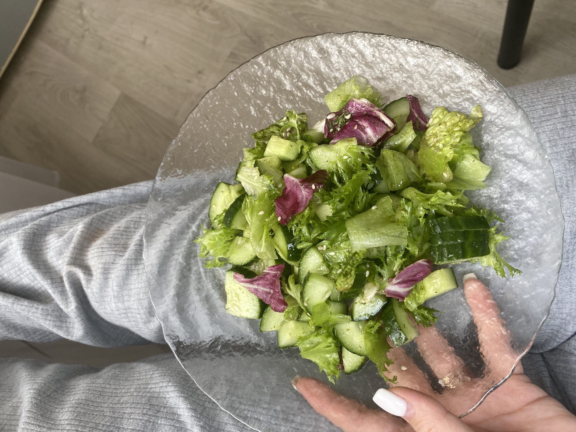 A person wearing gray sweatpants is holding a clear, textured glass plate of salad. The salad contains lettuce, cucumber, and red cabbage. The location is Chaikovsky, Russia. The plate is supported by the person's left hand. The right hand is visible supporting the plate.