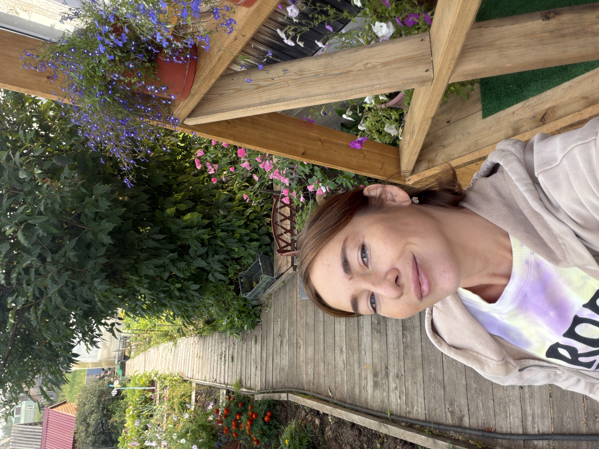 A woman is taking a selfie in a garden in Zheleznodorozhnyi, Russia. She is in the foreground, with a wooden path stretching out behind her and leading into the garden. The garden is filled with various plants and flowers, including pink roses and purple lobelia. A wooden structure, possibly part of a bench or swing, is to her right. In the background, there are houses and trees, typical of a suburban or rural setting. The woman is wearing a light-colored hooded sweatshirt over a tie-dye t-shirt. The sun appears to be setting, casting a warm glow on the scene.