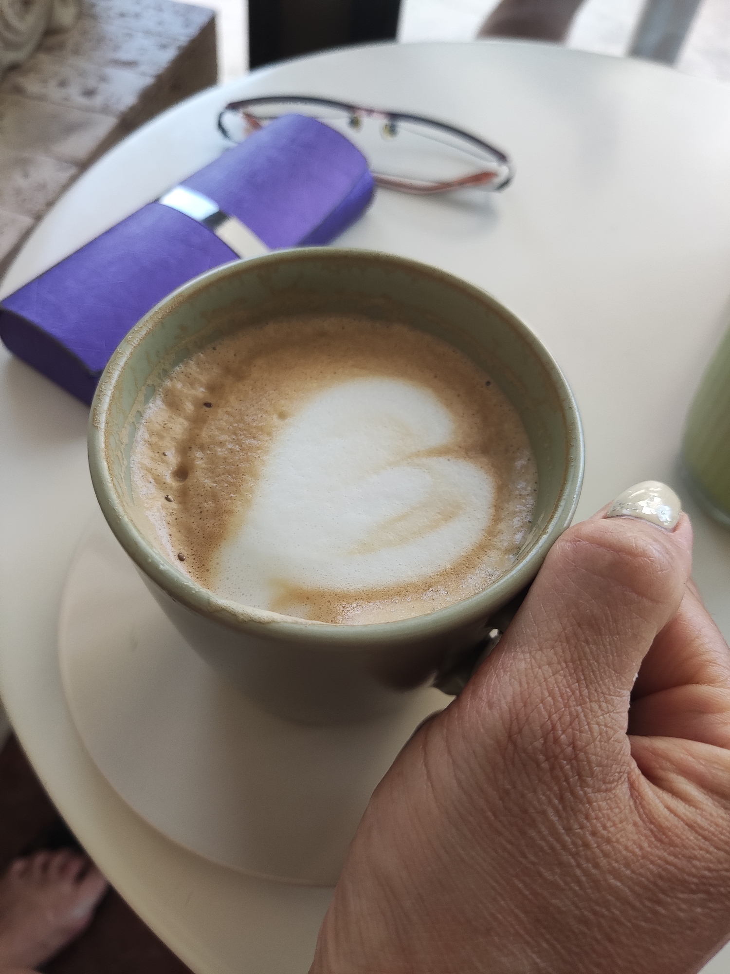 A close-up, eye-level shot shows a hand holding a cup of latte with a heart-shaped foam art, placed on a saucer on a white table. A purple eyeglasses case and a pair of glasses with red accents are on the table behind the cup. In the lower left corner, a person's bare feet are visible, suggesting they are sitting at the table. The scene takes place outdoors, likely on a terrace or patio, with a blurred background indicating a cafe setting in the city of Bar, Montenegro.