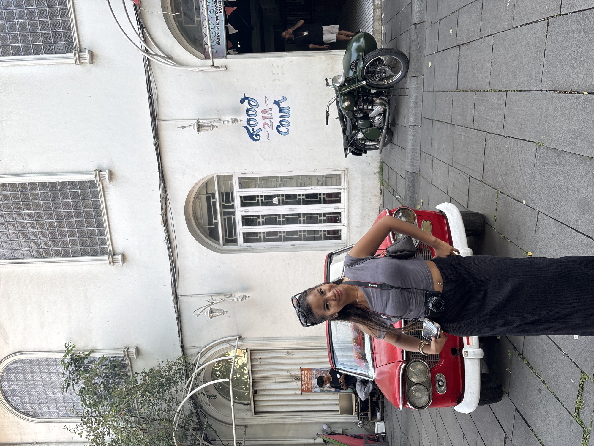 A young woman poses next to a vintage red car in front of a building with the text "Food 21A Court" painted on the wall. A motorcycle with a sidecar is parked nearby. The location is West Jakarta, Indonesia, based on the text "JASA FOTO KOTA TUA" and "ANEKA KULINER MAK KOTA" visible in the image. The woman is wearing a gray top, black pants, and has a camera around her neck.