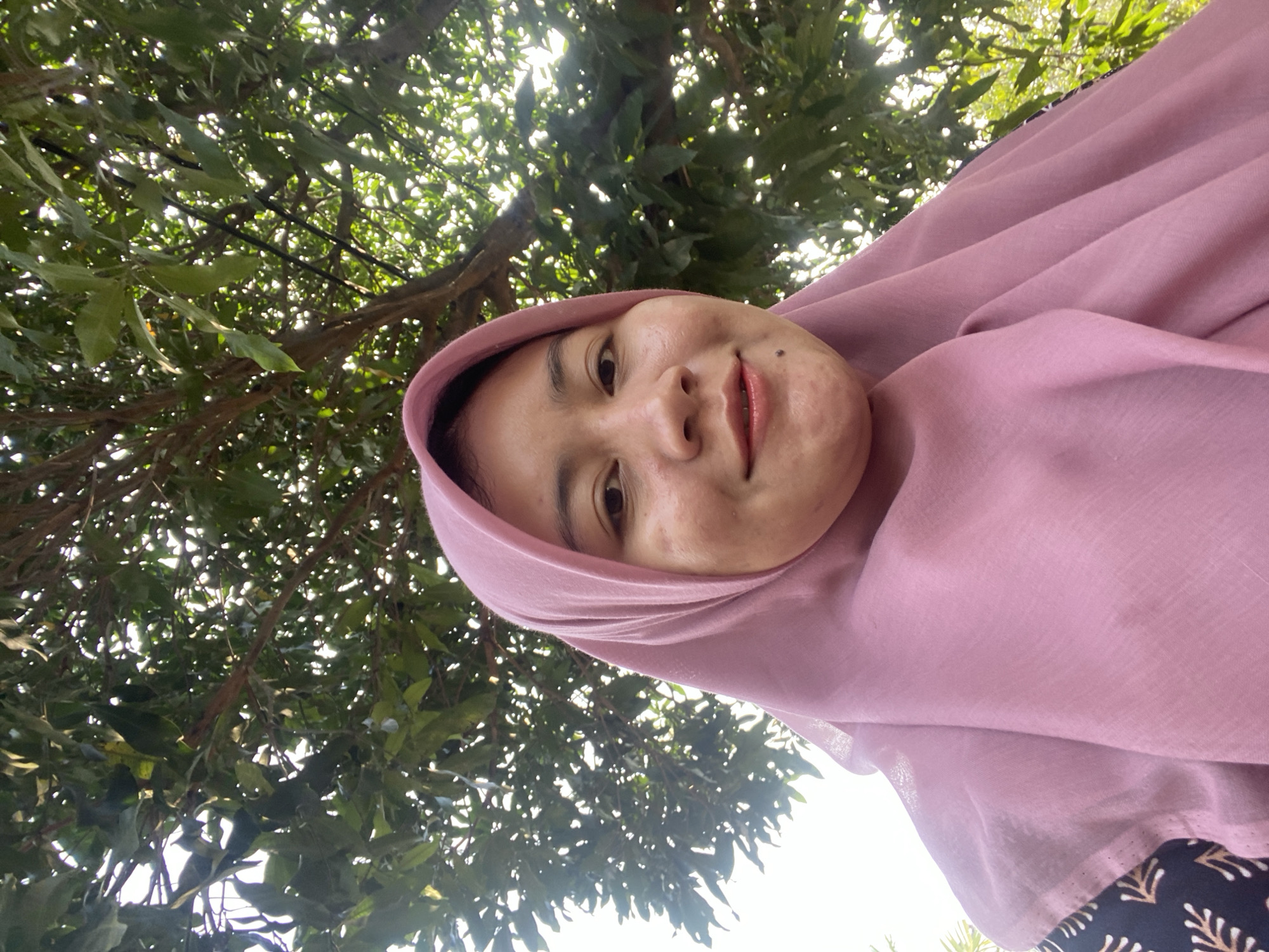 An Indonesian woman wearing a pink hijab and a patterned top smiles at the camera, framed by the dappled sunlight filtering through green leaves of a tree. The scene appears to be outdoors, possibly in a residential area or park in Banjarbaru, Indonesia, with the bright sky visible through the foliage.