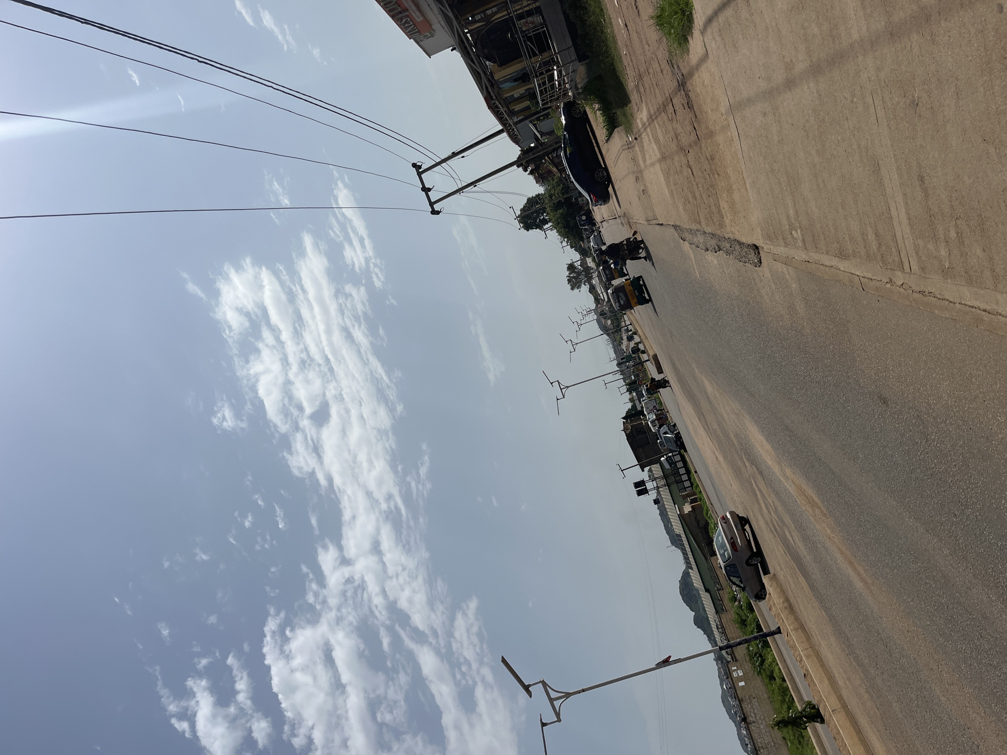 A tilted, eye-level, full shot captures a street scene in Bukuru, Nigeria, under a partly cloudy sky with visible power lines overhead. The paved road runs diagonally from the bottom left to the upper right. Multiple vehicles are present, including a beige sedan in the middle ground on the left, a black sedan on the right side of the road, a yellow auto-rickshaw further down the road on the left, and several motorcycles with riders. A pedestrian can be seen on the right side of the road. The environment includes a sidewalk on the right, lined with buildings and vegetation. Streetlights are visible along the road, and in the background, there are hills and more distant buildings. The image appears to be taken during daylight hours.