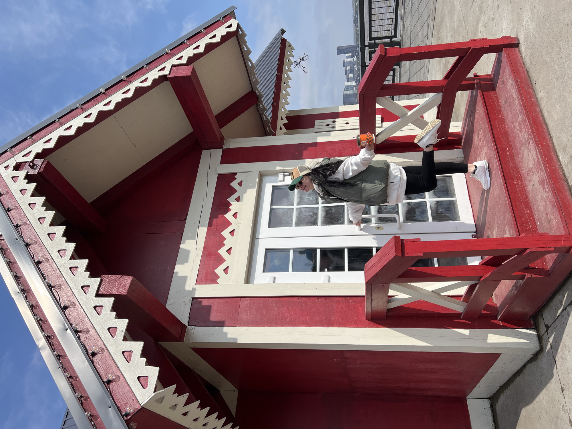 The image shows a woman standing sideways on the roof of a small, red and white building. She is wearing a white sweatshirt, black pants, white sneakers, and a green vest, and a green baseball cap with a brown brim. She is holding a small orange object in her right hand. The building has a window with multiple panes and decorative trim along the roofline. The sky is visible in the background. The building appears to be part of an attraction, and based on the architecture (particularly the decorative trim and color scheme), it's likely in Kazan, Russia. Tall buildings can be seen in the background.