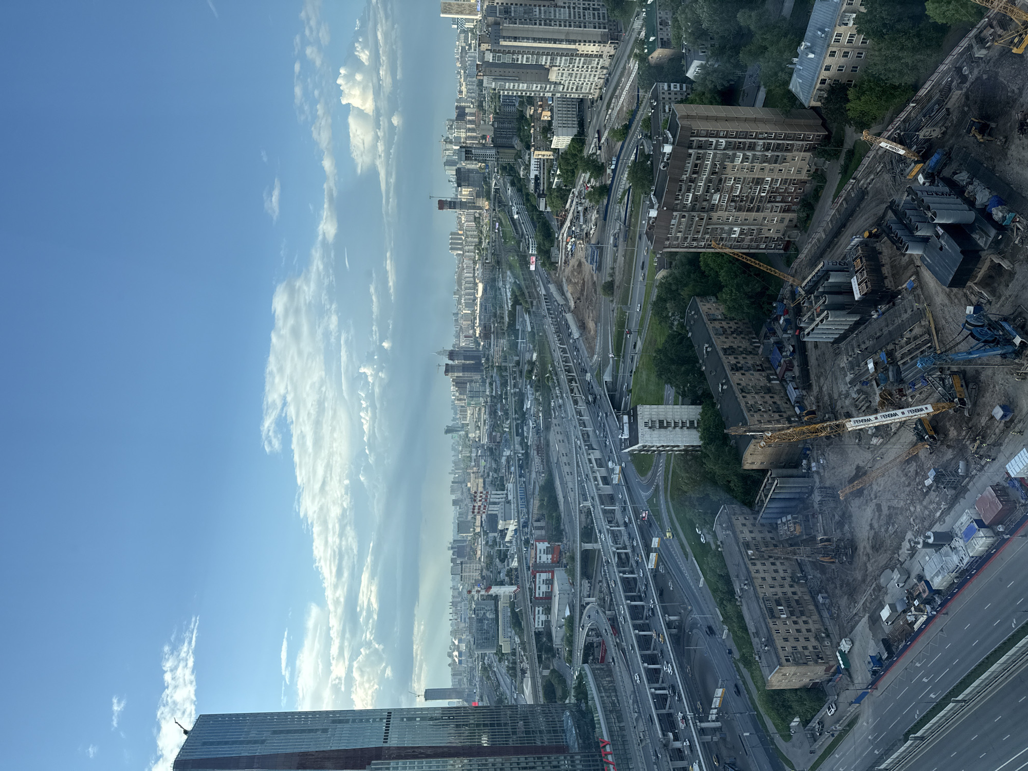 The image is a high-angle, daytime view of Khimki, Russia. The sky is blue with white clouds. A dense cityscape stretches from the foreground to the horizon, featuring buildings of various heights and architectural styles. Major infrastructure elements are visible, including highways, overpasses, and a network of roads carrying vehicular traffic. A construction site with cranes and construction materials is present, possibly indicating ongoing urban development. The image shows high-rise buildings and a wide range of buildings with various architectural details.