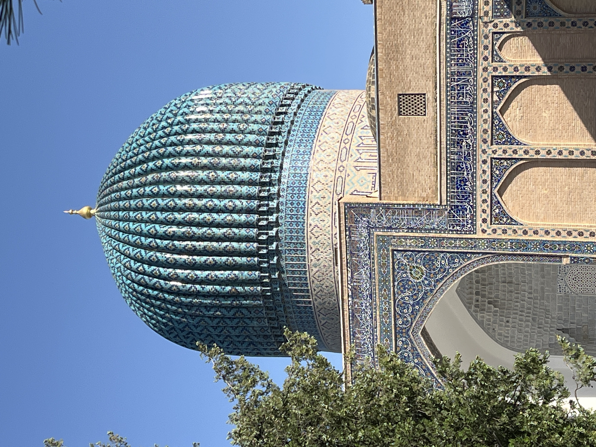 The image displays a close-up view of the azure dome of a mosque, set against a clear, bright blue sky. The dome is intricately decorated with turquoise and blue tiles arranged in a patterned, ribbed structure, culminating in a golden finial. To the right of the dome, a section of the mosque's building is visible, constructed from light brown bricks. This section is adorned with elaborate blue and white tilework featuring Arabic calligraphy and geometric patterns. The ornate tilework also frames an arched entryway, revealing a glimpse of the mosque's interior. Lush green foliage from trees partially obscures the lower portion of the structure, suggesting an outdoor setting. The lighting indicates it is daytime, likely a sunny day, given the clear sky and the shadows cast on the building. The architectural style, particularly the dome and the detailed tilework, strongly suggests a location in Uzbekistan, with Samarkand being a prominent center for such historical and religious architecture. No people are visible in the image.