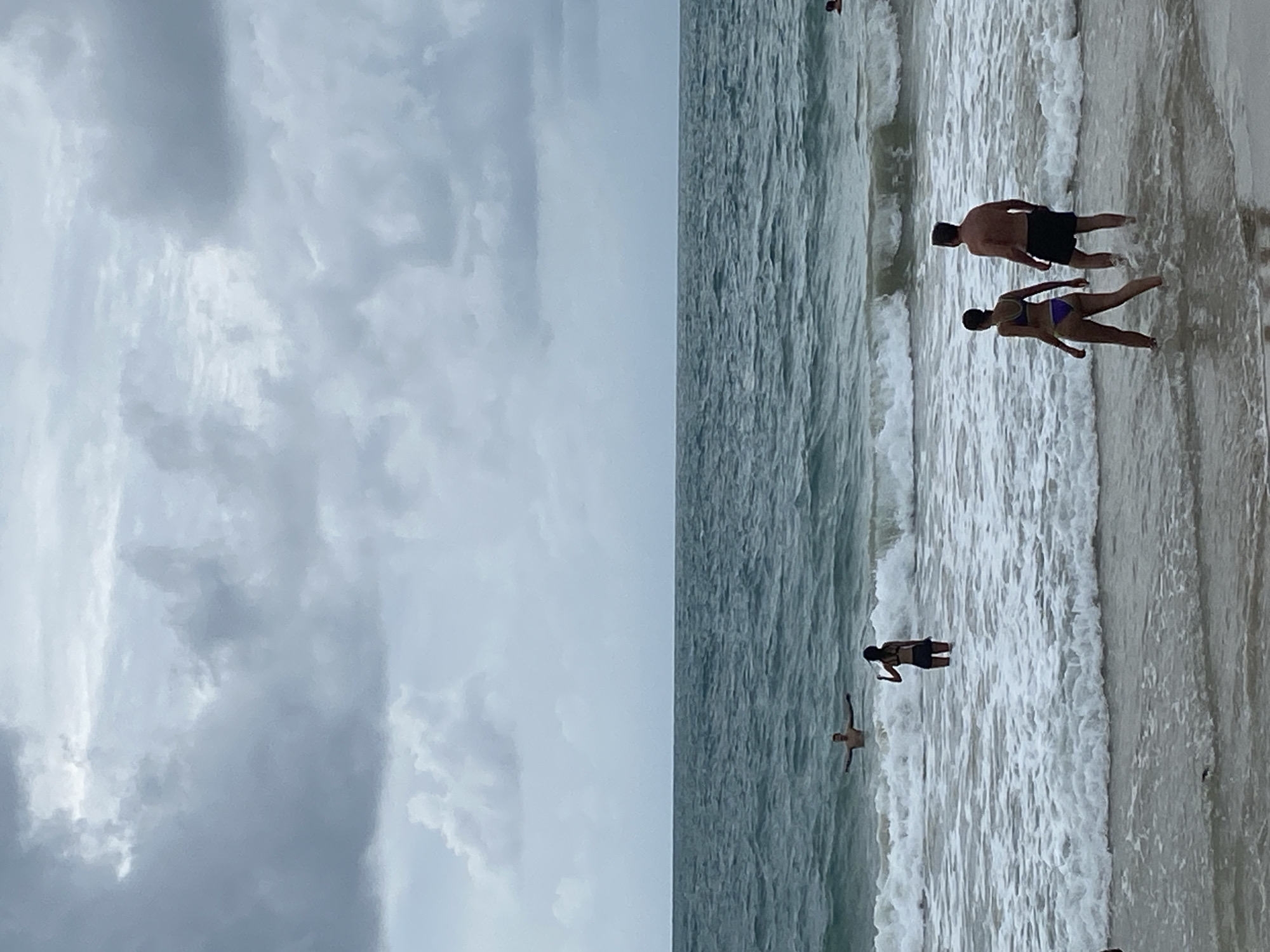 The image depicts a beach scene in Choeng Thale, Thailand. A wide expanse of cloudy sky fills the left portion of the image. The right side shows a beach with several individuals wading and playing in the ocean. A couple stands near the shoreline, and two other individuals are further out in the water. The tide is coming in, with foamy waves breaking near the shore.