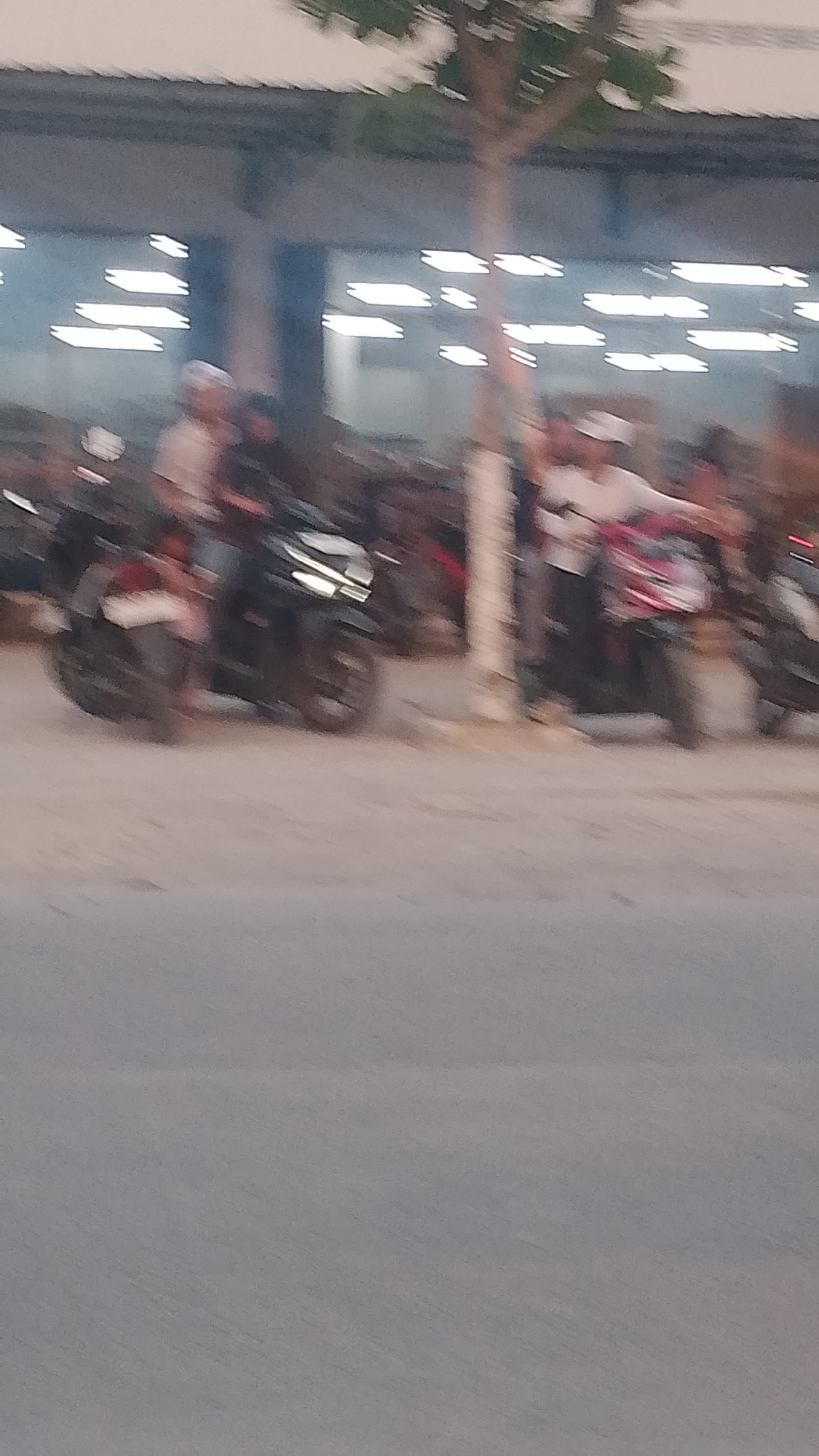 The image shows a street scene in Karanglangu, Indonesia, with several motorcycles parked and people milling around. The lighting suggests it is dusk or early evening. A covered structure, possibly a building or awning, is visible in the background, with rows of bright lights. A tree with sparse foliage stands near the center of the image. The ground appears to be paved. The people in the image are blurred due to motion, making it difficult to discern specific features or actions. However, some individuals appear to be standing near or on motorcycles. The overall impression is of a public gathering or transit area.