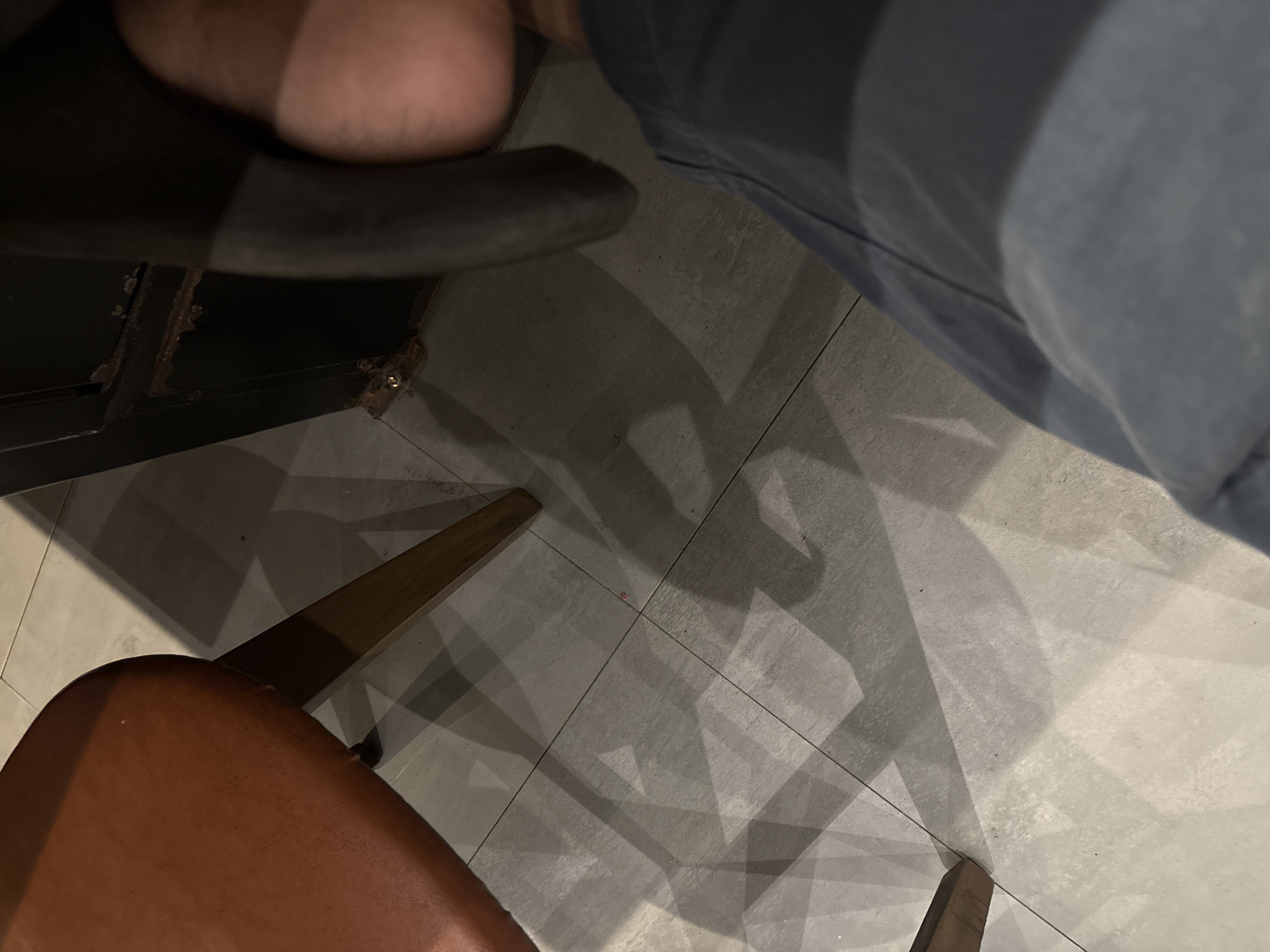 A close-up, high-angle shot shows a person's legs and feet in dark pants, with a shadow of what appears to be a chair cast across a tiled floor. The chair, partially visible in the upper left, has a brown, padded seat and wooden legs. The floor tiles are large and light gray. To the right, a dark metal structure with visible rust and a small screw is present. The lighting suggests an indoor setting with a light source creating distinct shadows. The scene is devoid of any specific landmarks or clues to pinpoint its location in Cirebon, Indonesia.