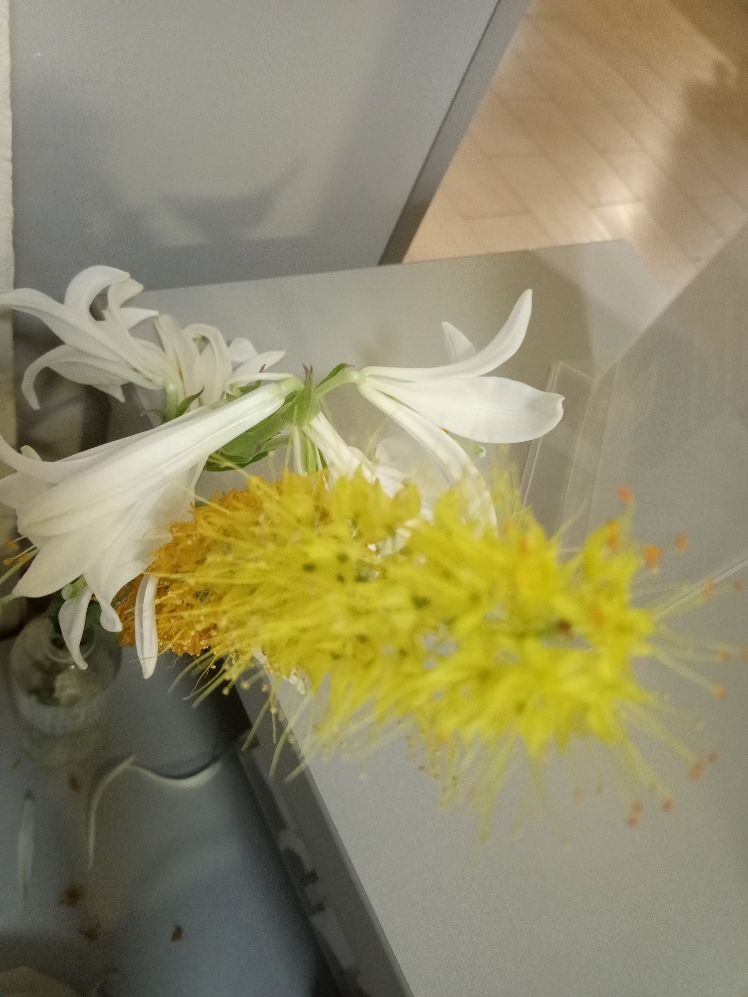 A floral arrangement with white lilies and yellow flowers is placed on a gray surface, possibly a table or counter. The background is out of focus but appears to be a wall and a wooden floor. The lighting suggests an indoor environment.