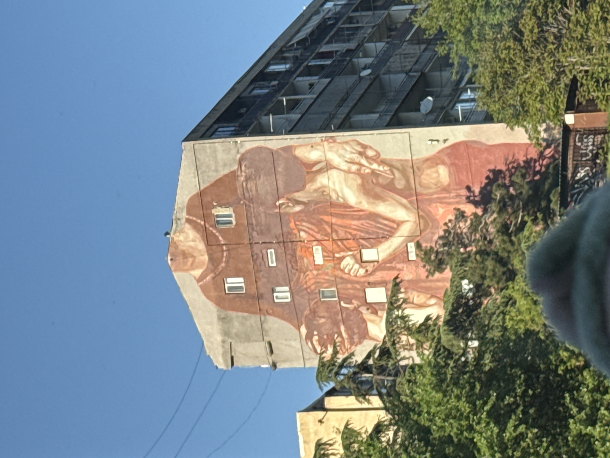 The image shows a high-rise building in Tbilisi, Georgia. The building has a mural on its side depicting figures that seem reminiscent of Renaissance art. The mural is integrated into the building's architecture, using the windows as part of the artwork. Trees obscure the lower part of the mural and some of the lower building levels. The sky above is clear and blue.