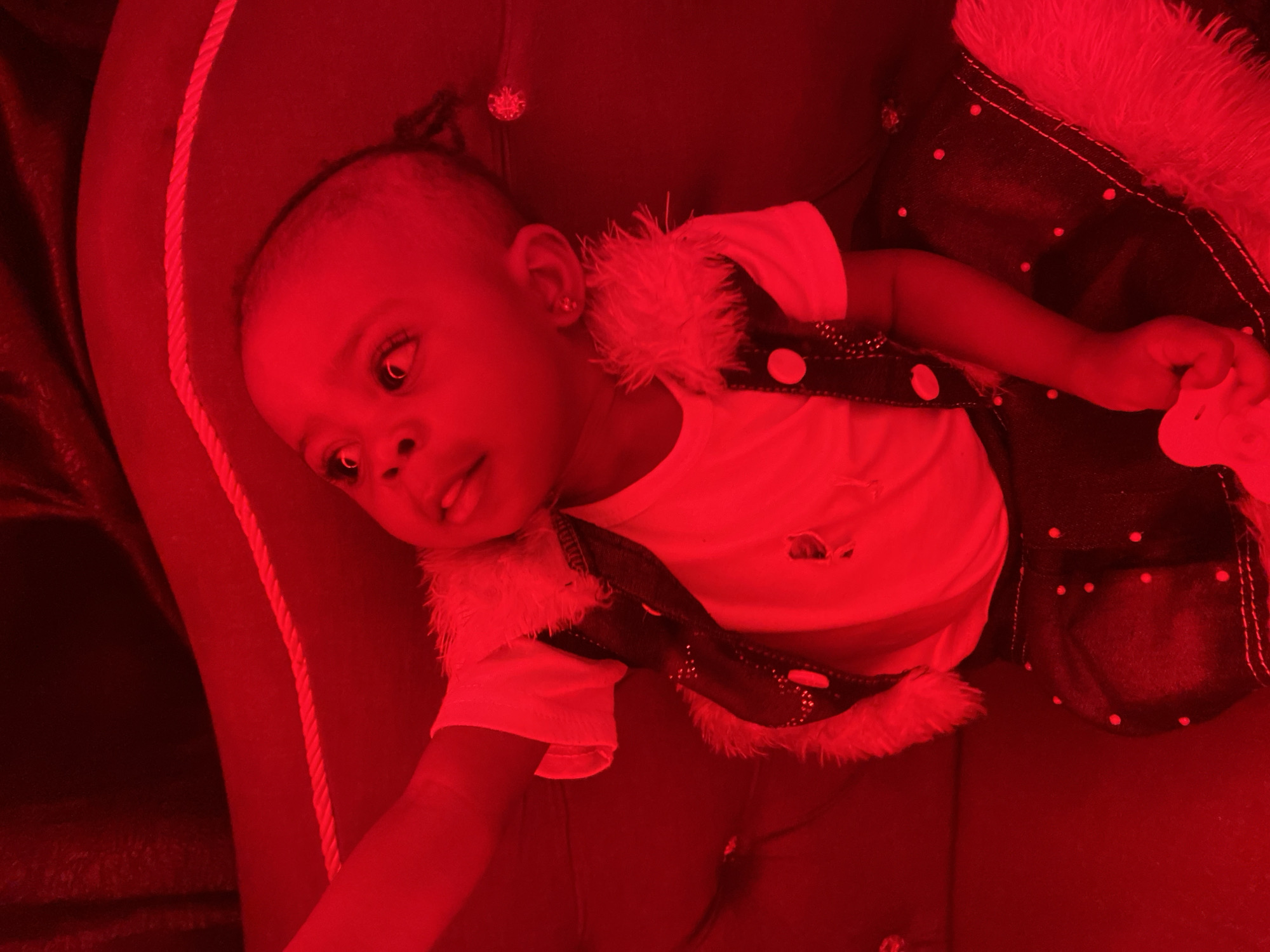 The image shows a close-up view of a young child, likely a toddler, sitting on a red couch. The child is wearing a white t-shirt, a dark vest with fur trim and red polka dots, and a matching dark skirt with fur trim. The child has dark skin, dark eyes, and dark hair styled in a small ponytail. The child is looking upwards and to the left with an expressive gaze. The overall lighting of the image is a strong red hue, which casts a deep crimson tone over the entire scene. The couch appears to be upholstered in a textured material, with a decorative braided rope element visible in the background. The setting is indoor, with soft lighting contributing to the red tint. There is no indication of the specific location within Abakaliki, Nigeria.