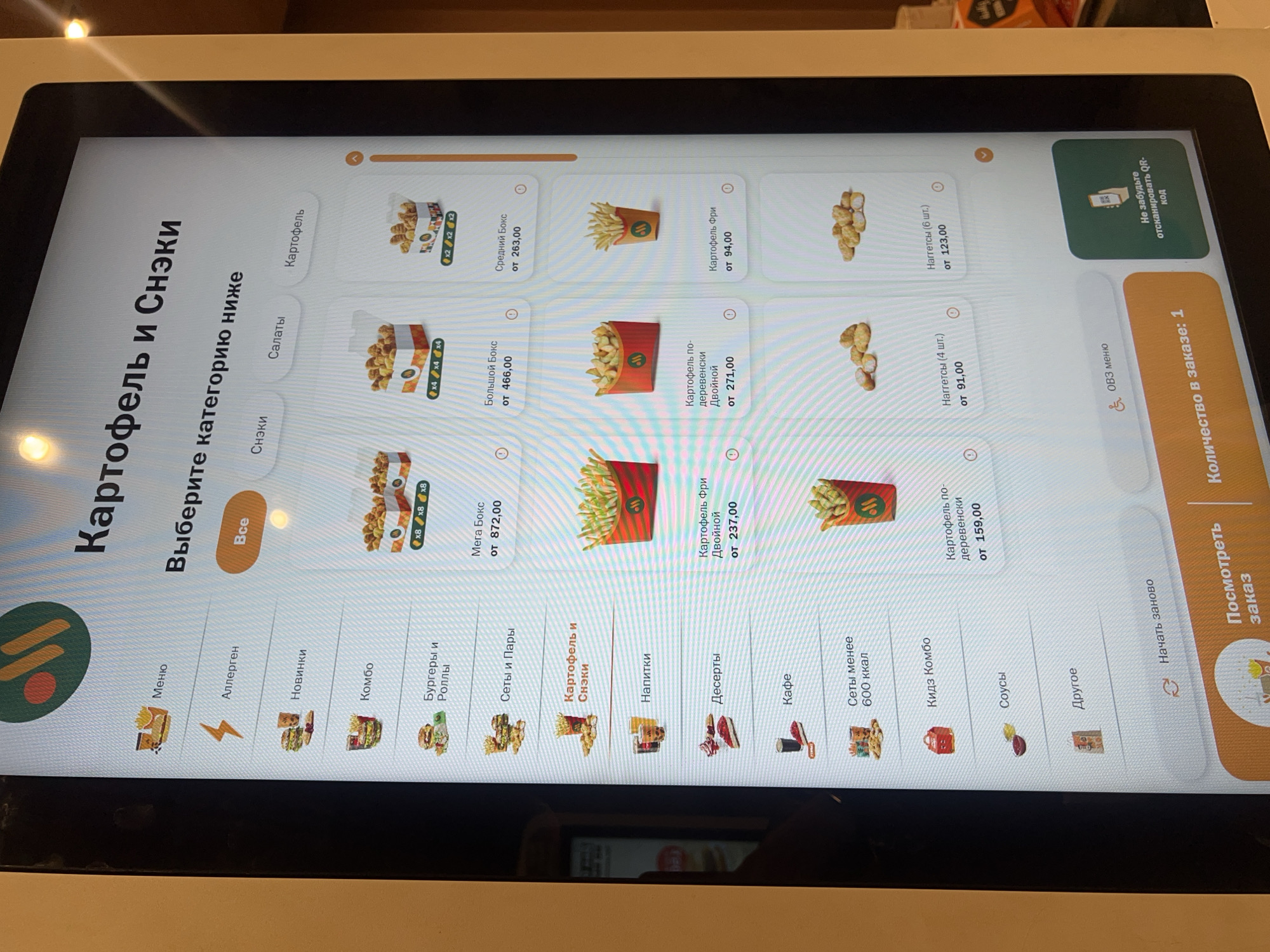 The image shows a digital self-ordering kiosk, likely inside a McDonald's restaurant. The screen displays a menu with various food categories, including "Potatoes and Snacks" which is selected. Under this category, different potato-based items such as fries, and "boxes" are shown along with their prices. Other menu categories like "Burgers", "Drinks", and "Desserts" are also visible. The bottom of the screen includes further options such as "sauces," "kid's combo," and "coffee." A progress bar on the right indicates "Number in order: 1". A QR code for scanning is displayed on the upper right. The kiosk is showing a Russian language menu.
