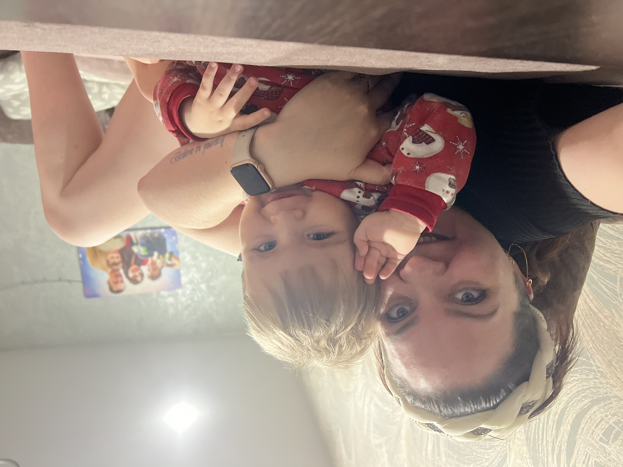 A selfie shows a woman and a young child. The child wears a red Christmas-themed outfit. The woman has brown hair and is wearing a patterned headband. She is wearing a smart watch on her left arm. A family photo is visible on the wall in the background. The environment appears to be a bedroom or living room. It is not possible to determine from the image if this photo was taken in Noviy, Russia.