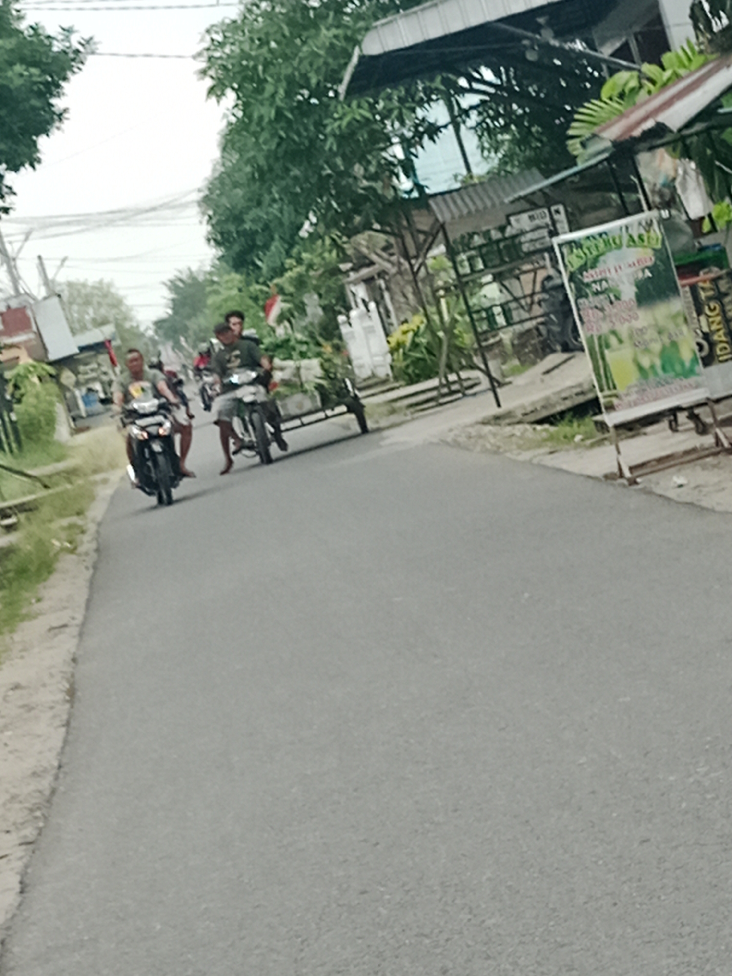In Binjai, Indonesia, on a paved road with a dirt shoulder, two men are riding motorcycles side-by-side. The man on the left is on a black motorcycle, and the man on the right is on a darker colored motorcycle. Both are wearing casual clothing. Behind the man on the right, a small cart is attached to his motorcycle. Trees line the right side of the road, and a small stall or building is visible. A sign with green text and graphics stands near the stall. The sky is overcast.