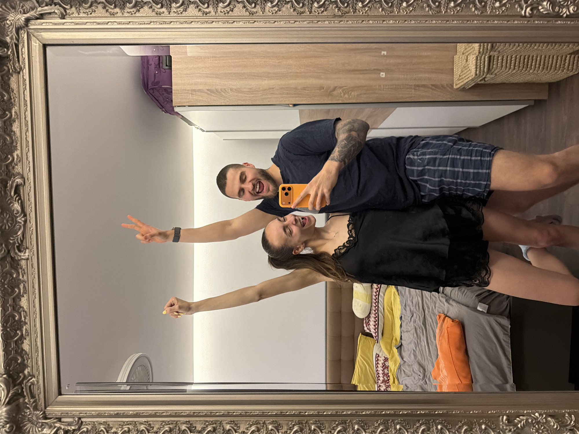 A couple is taking a selfie in a mirror with an ornate frame. The man is wearing a dark blue t-shirt and plaid shorts, holding an orange smartphone. The woman is in a black top. The background reflection shows a bedroom with a bed covered in various colored pillows and a closet in the upper portion. A partially visible fan is in the lower corner. There is no definitive information in the image to locate this within Sofia, Bulgaria.