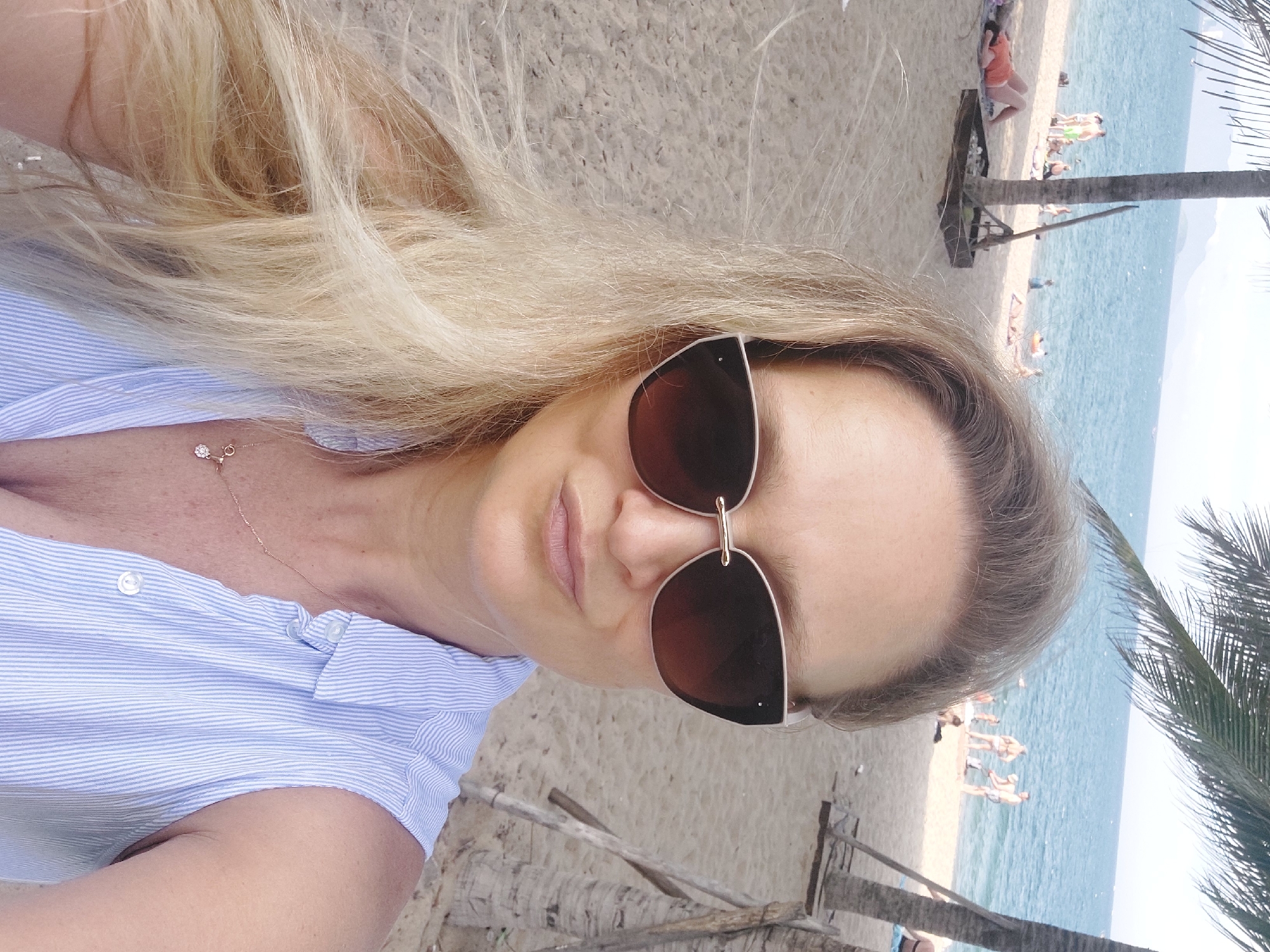 A blonde woman with sunglasses is taking a selfie on a beach in Nha Trang Ward, Vietnam. She is wearing a blue and white striped sleeveless shirt and a delicate necklace with a pendant. The beach is sandy, with palm trees in the background. The ocean is visible, with several people sunbathing and swimming. The weather appears to be sunny and warm.