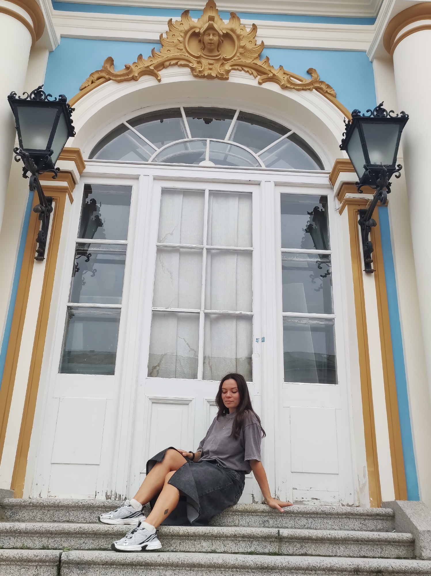 A young woman with long dark hair is sitting on the granite steps in front of a white door. She is wearing a gray t-shirt and a dark denim skirt. Her eyes are closed and she is leaning her right arm on the step. She is wearing silver and black sneakers. The door has multiple glass panes and is framed by columns painted white, yellow, and blue. There are decorative black lanterns on either side of the door. The building appears to be a palace or a grand building. This is likely the Catherine Palace in Pushkin, Russia, known for its ornate architecture and blue and gold accents.