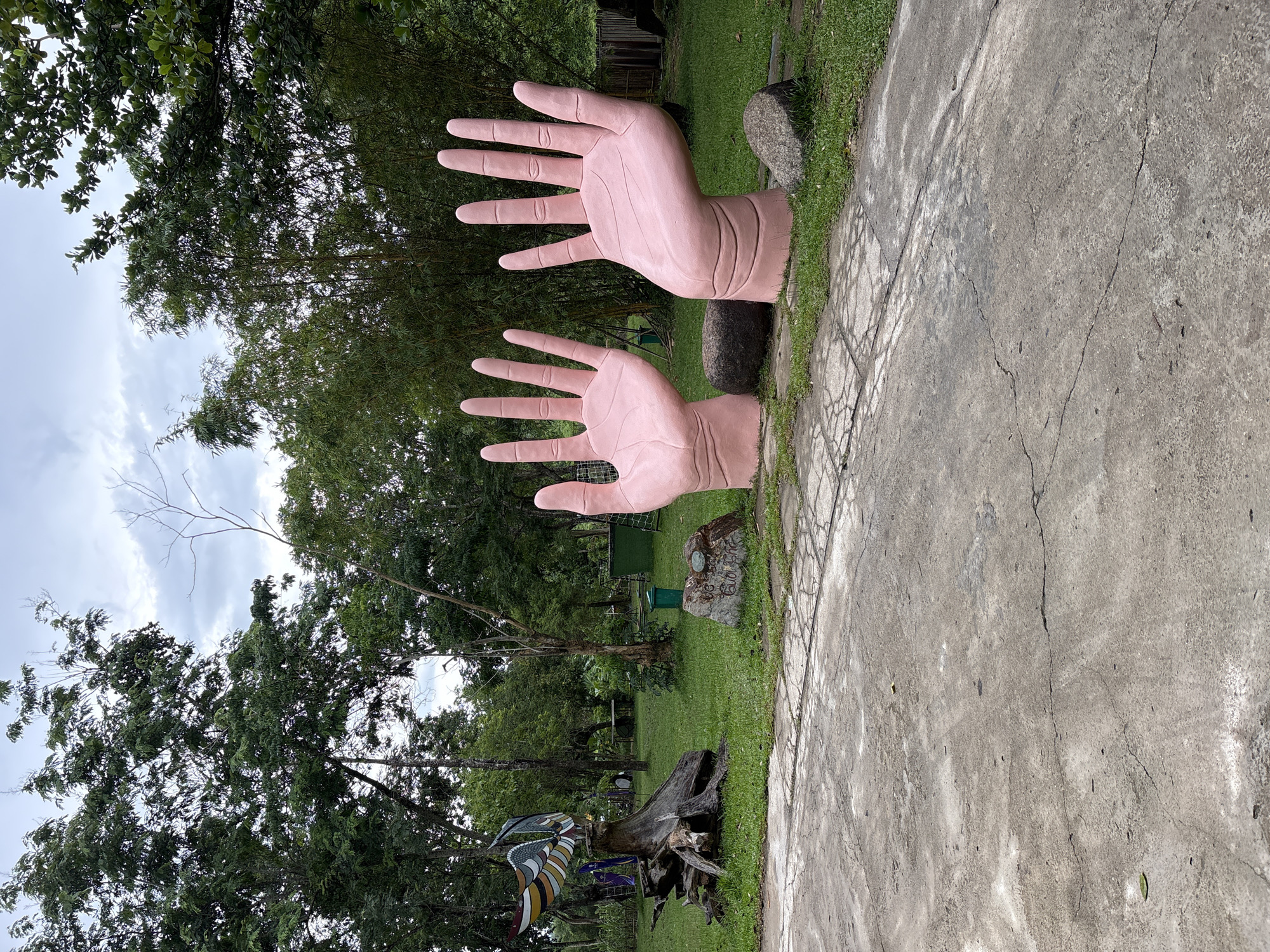 The image depicts an outdoor scene in what appears to be a park or garden area. In the foreground, a cracked concrete pathway leads towards a grassy area. Two large, pink, sculpted hands, palms facing upward, emerge from the ground. The left hand is positioned slightly behind and to the left of the right hand. Between the hands and to the right of the left hand are rounded, dark-colored rocks. Further into the background, on the left side of the frame, a weathered tree stump serves as a base for a colorful, abstract sculpture resembling a bird with outstretched wings. Behind the stump, several trees and green foliage are visible. A green trash bin is situated in the middle ground, to the left of the right sculpted hand. In the background, dense green trees and bamboo stalks create a natural backdrop under a cloudy sky. The overall impression is one of an art installation set within a natural environment. There are no people visible in the image.