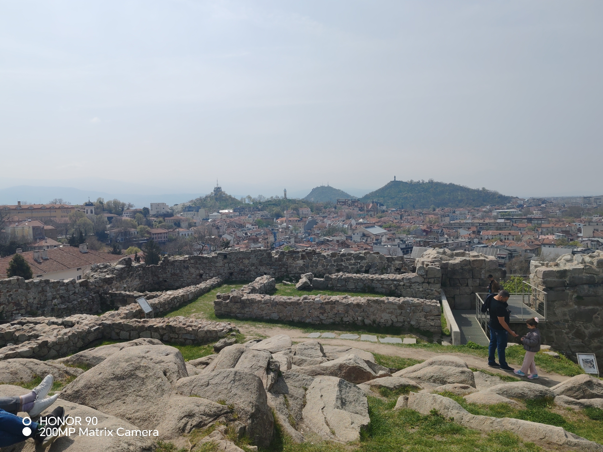 The image depicts a panoramic view of Plovdiv, Bulgaria, from the Nebet Tepe ruins. Stone ruins are visible in the foreground, with a mixture of stone walls and large boulders covered in grass. In the midground, a cityscape of tightly packed buildings with red-tiled roofs spreads out towards three distant hills, each topped with a structure. A man and a young child are interacting near a modern metal railing, seemingly looking at something in the child's hand. The sky is overcast, and a brand identifier appears in the lower left corner of the image.