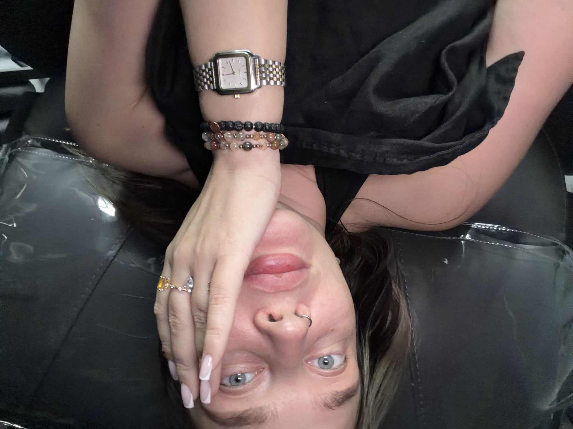 A female individual is positioned close to the camera, her face partially obscured by her hand. She has light skin, blue eyes, a nose ring, and light-colored nails. Her hair is visible around her face. On her wrist, she wears a watch and two beaded bracelets. Her hand has two rings. She is dressed in a black garment. The background is a dark, glossy surface, possibly a seat. The location is unknown and not apparent from the image.