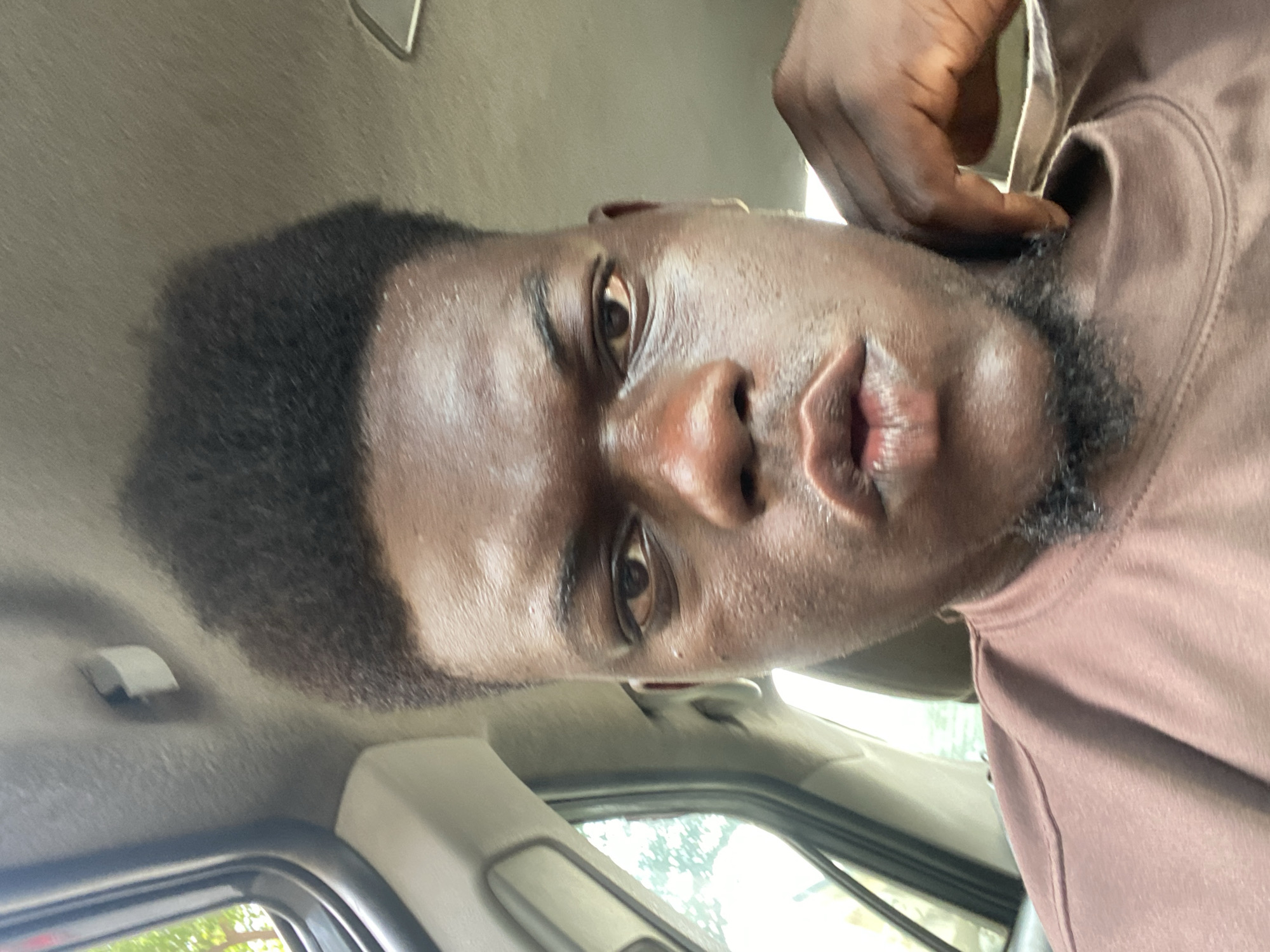 A close-up, slightly angled portrait of a Black man inside a vehicle. He has short, dark hair and a trimmed beard. His skin appears shiny. He is wearing a brown shirt. Part of the vehicle interior, including the ceiling and window frame, are visible in the background. The location appears to be a city street.