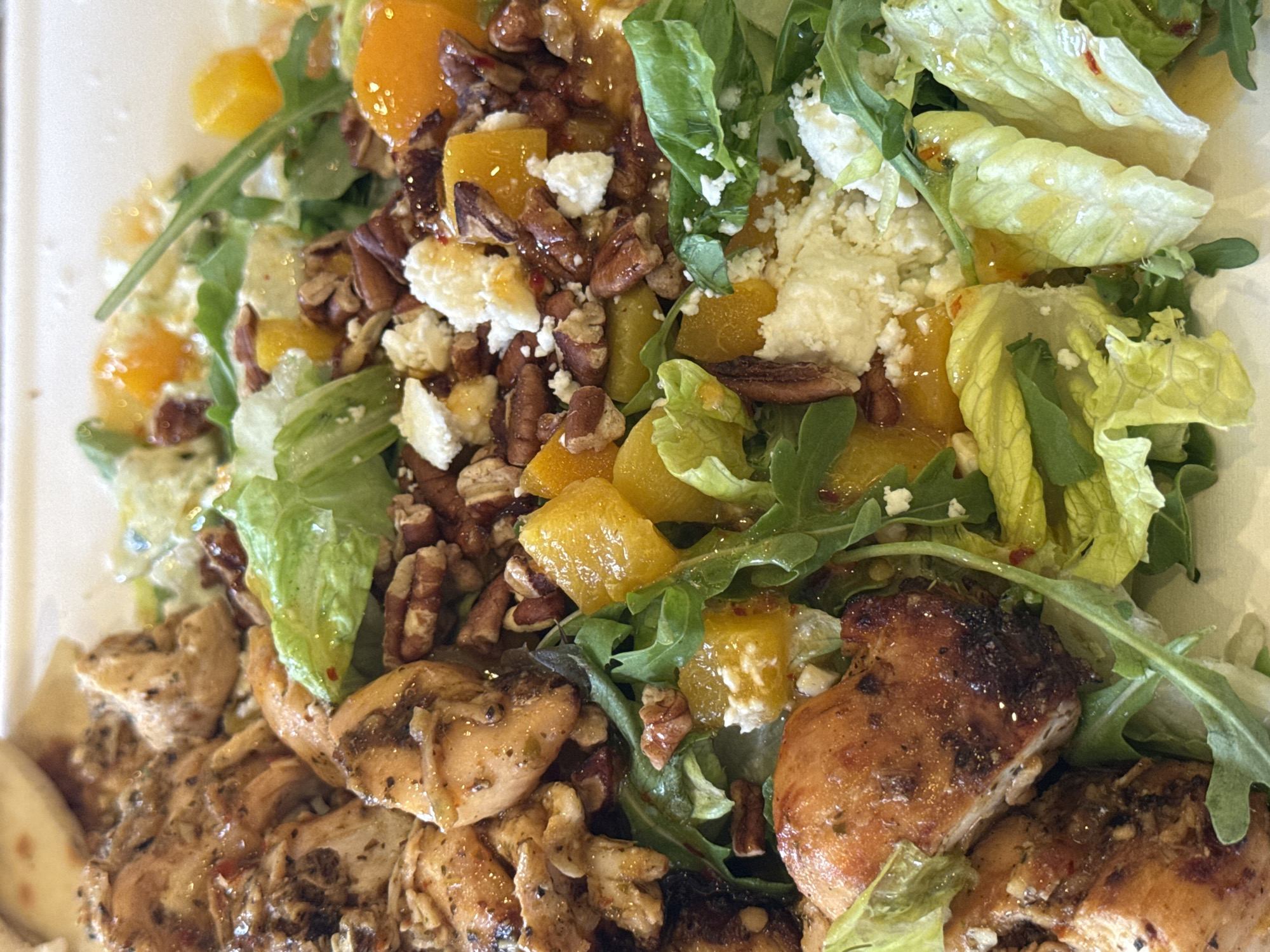 The image shows a close-up of a salad containing grilled chicken pieces, lettuce, arugula, cubed orange segments, pecans, and crumbled cheese. The salad appears to be dressed with a light vinaigrette. While the image does not offer sufficient context to confirm the location as Chula Vista, United States of America.