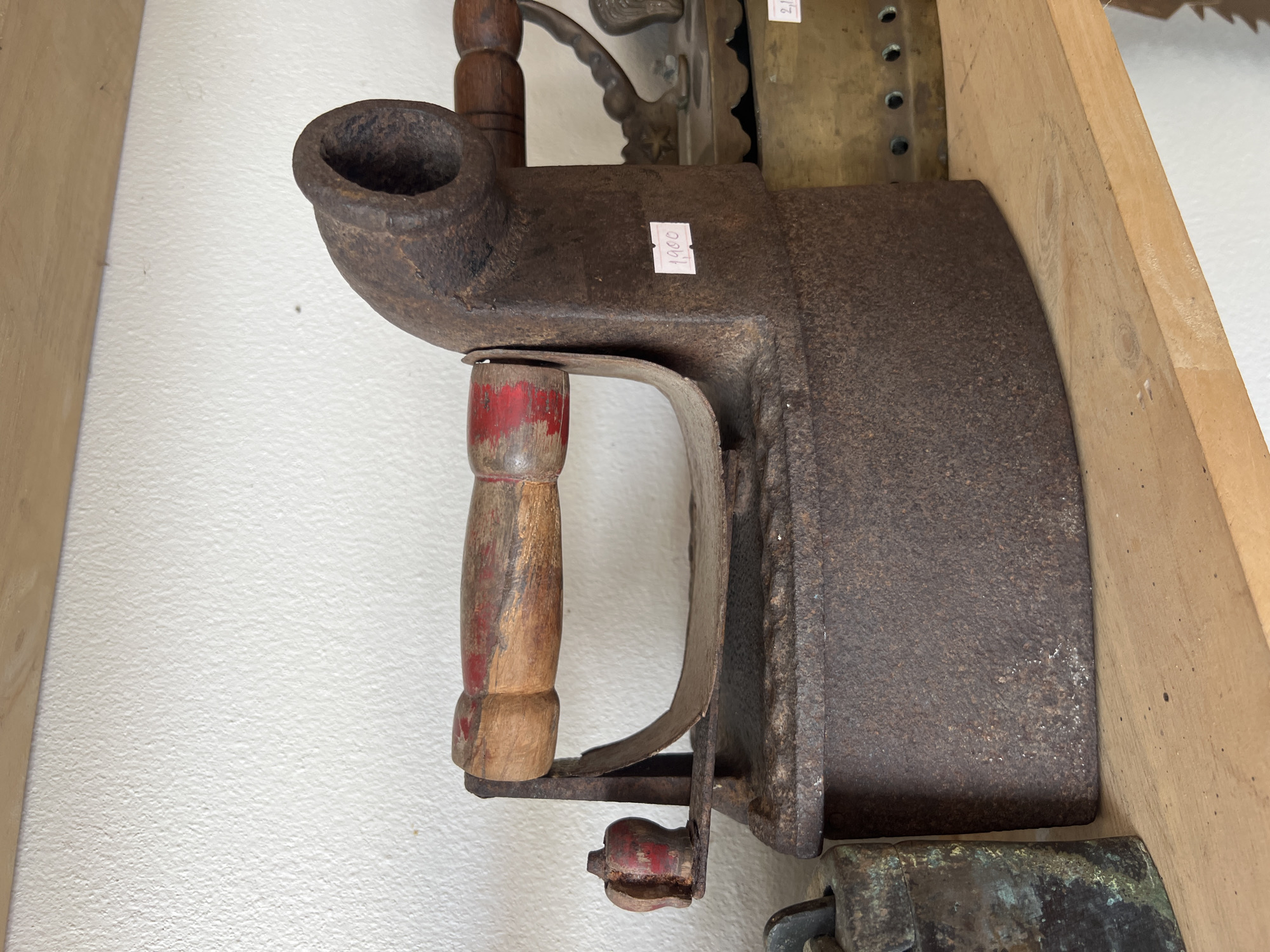 A close-up, eye-level shot shows vintage irons on a wooden shelf in what appears to be a shop or antique store. The primary subject is a large, heavily rusted cast iron flat iron with a wooden handle that has red paint remnants. A small, red-painted decorative knob is attached to the front of the iron. A price tag reading "1900" is affixed to the side of this iron. Behind it, partially visible, is a brass flat iron with decorative cutouts and another price tag indicating "2,000". To the left of the main iron is a dark, heavily oxidized metal object, possibly another antique item. The background is a textured, off-white wall. The overall lighting is soft and even, suggesting an indoor setting. The location is identified as Tambon Phraek Ha, Thailand.